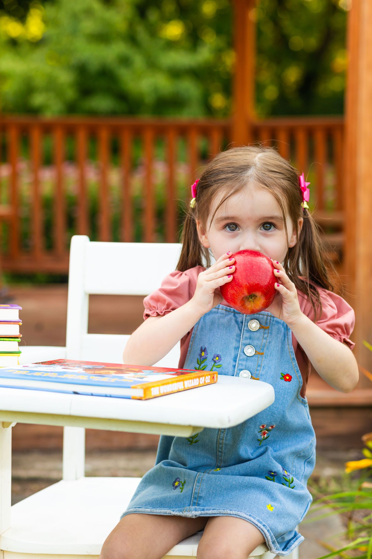 Child in denim overalls sitting at white table with books outdoors.