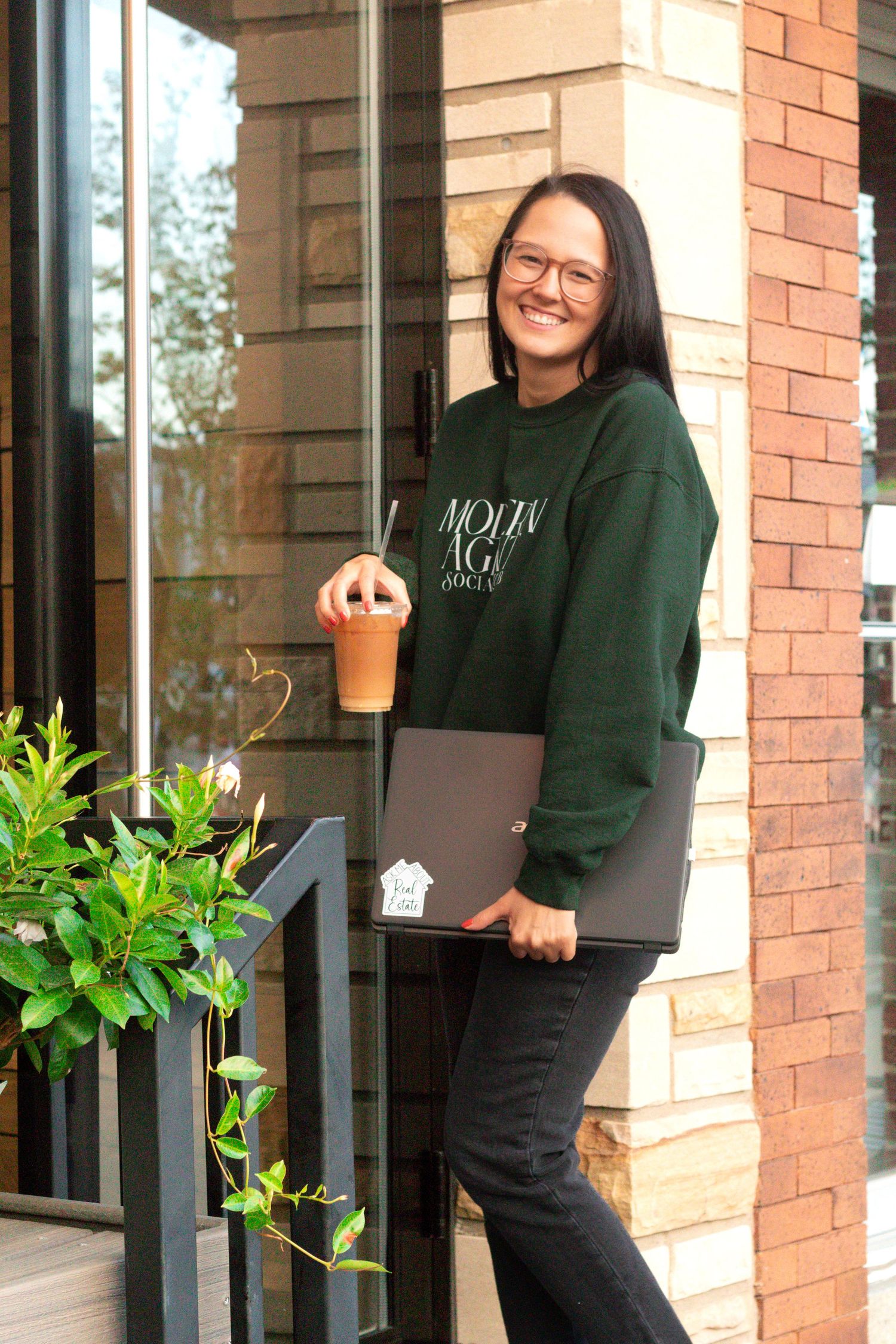 Person in green sweatshirt holding laptop and coffee outside brick building entrance.