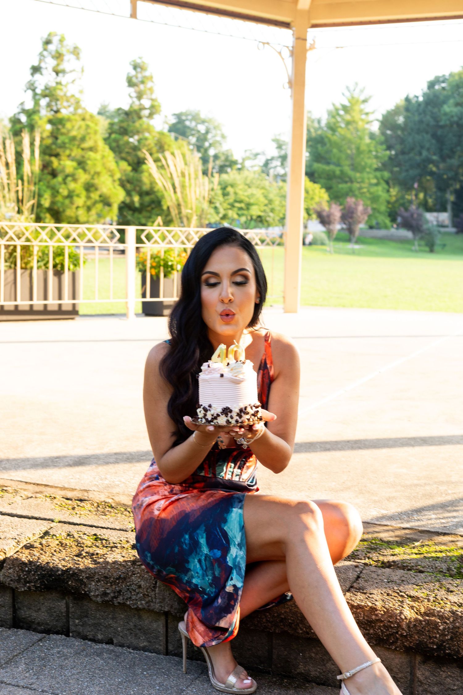 Woman in colorful dress blows out birthday candle on cupcake while sitting outdoors at sunset.