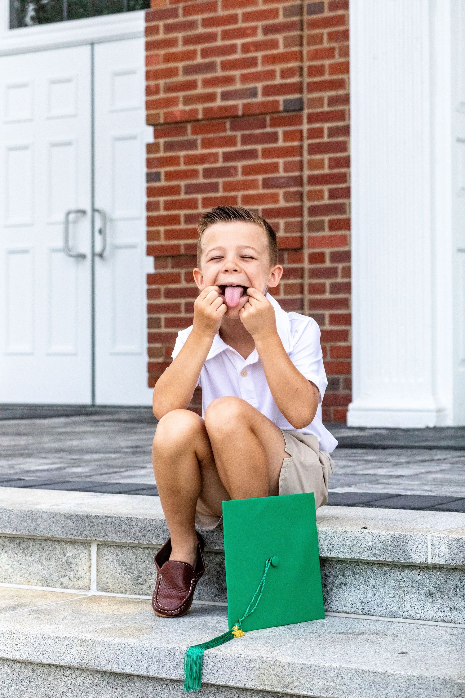 Young child in white shirt sits playfully on concrete steps with a green folder in front of brick building.