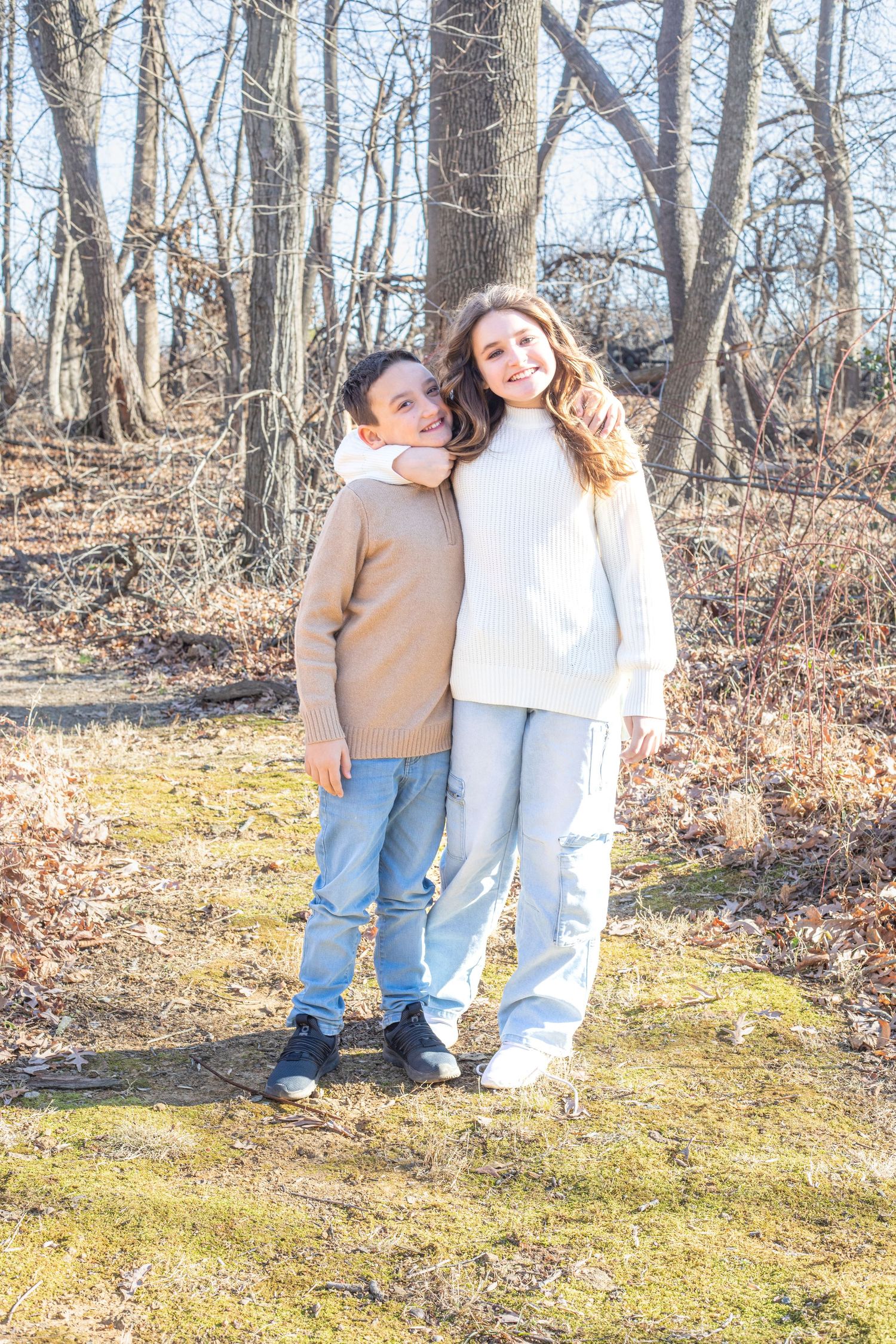 Two children in casual winter wear standing together in wooded area during winter.