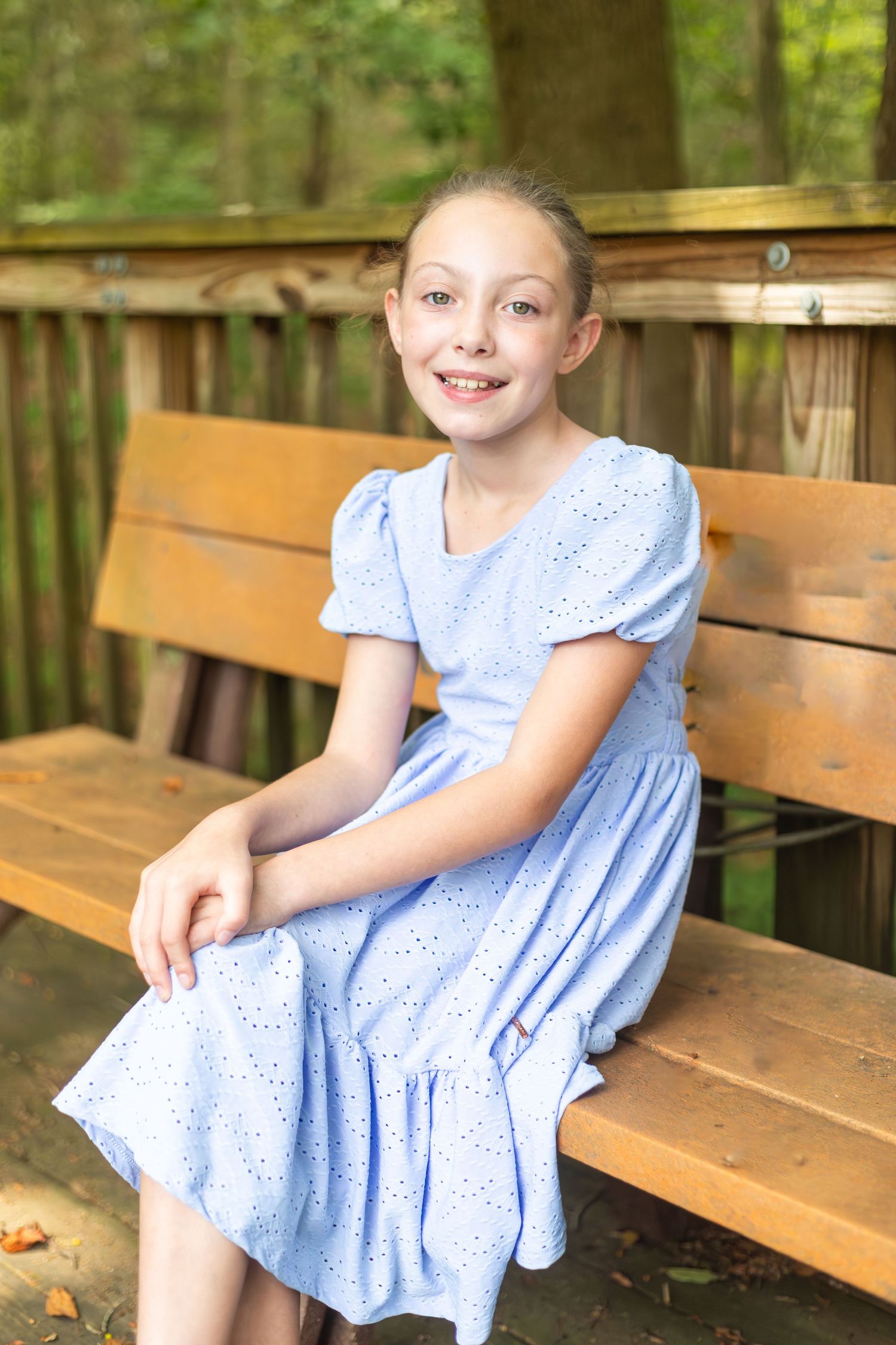 A child in a light blue eyelet dress sits on a wooden bench outdoors surrounded by greenery.