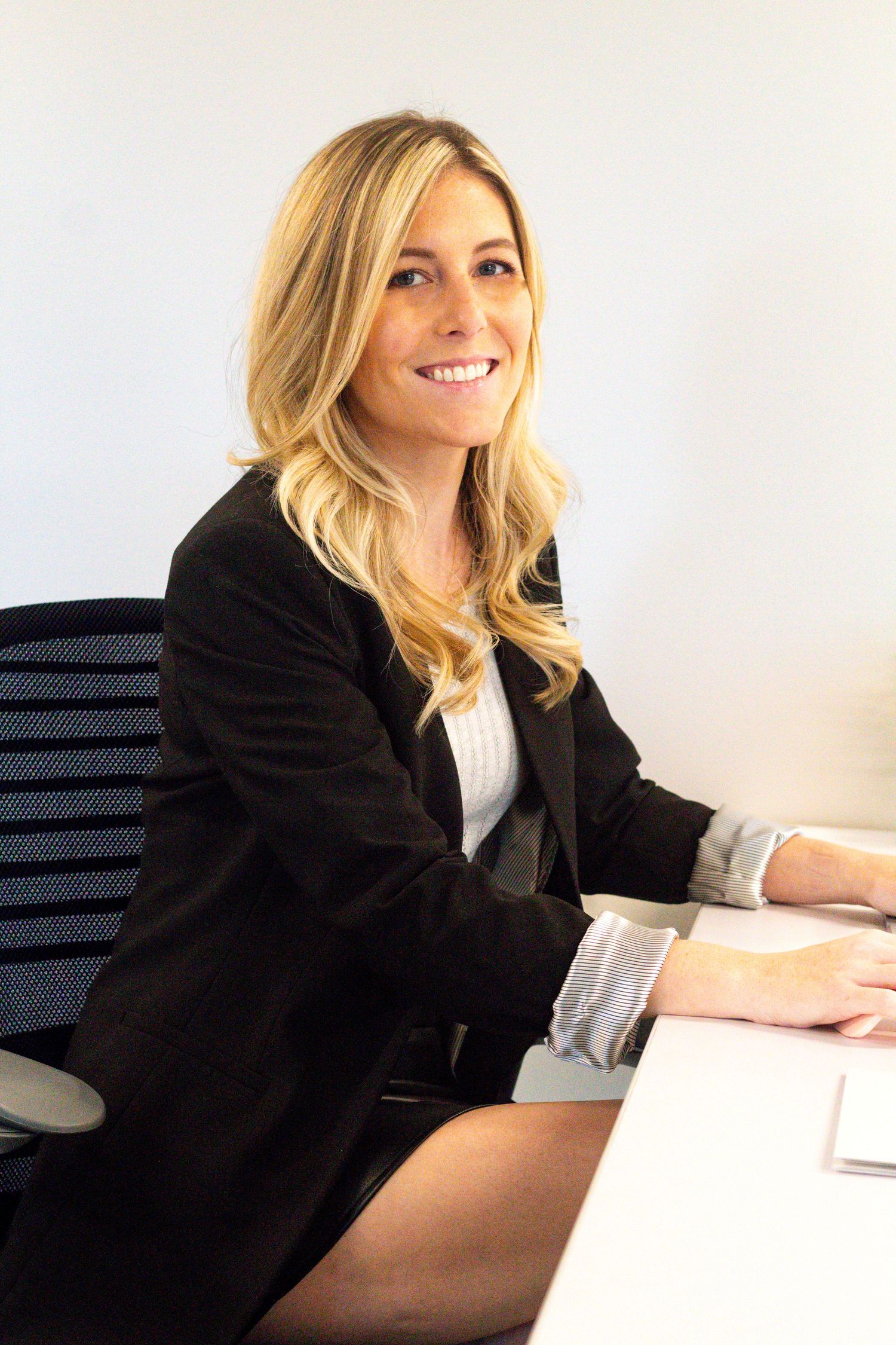Professional woman in black blazer sits at office desk with bright background.