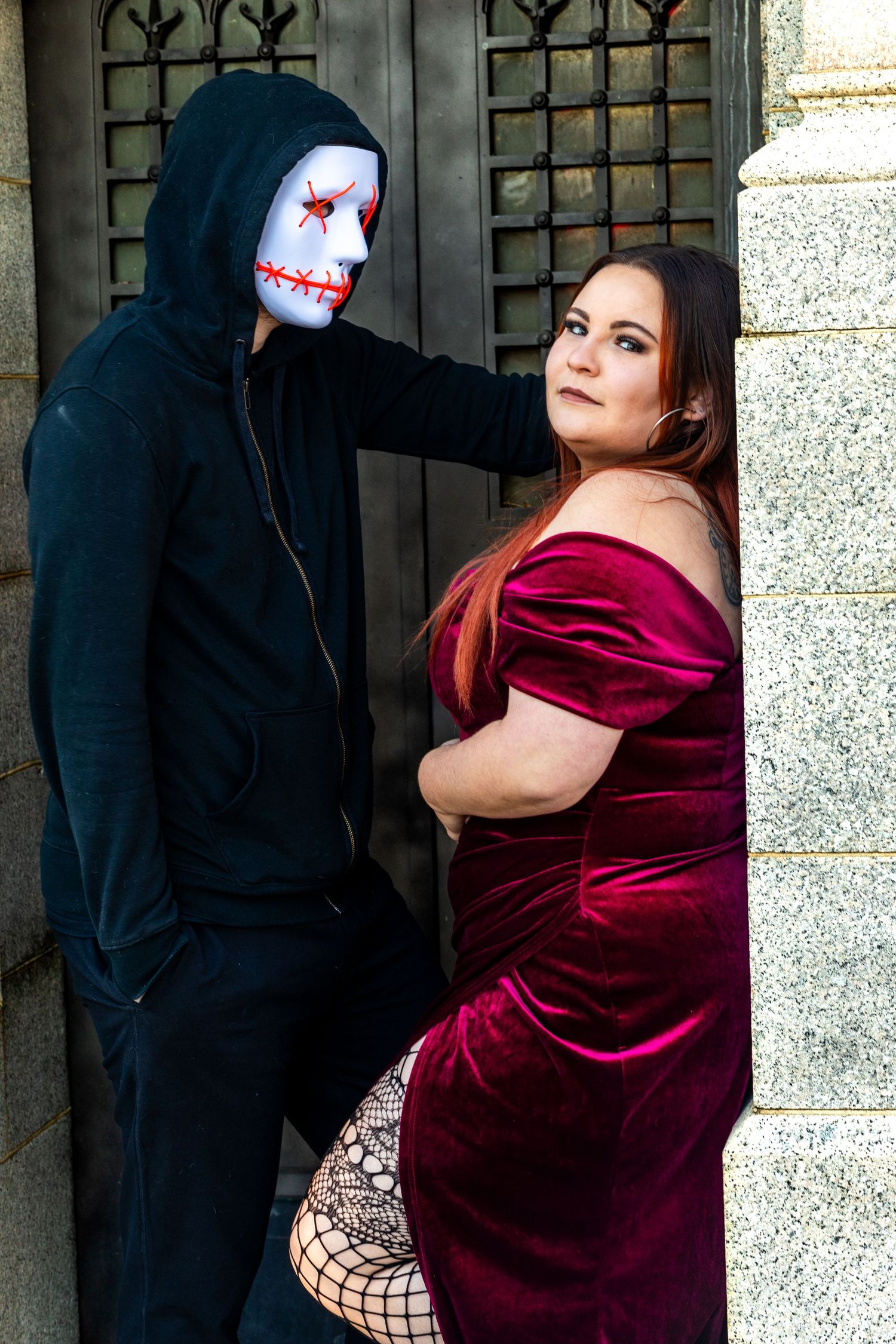 A haunting scene with a masked figure in black and someone in a red velvet dress against stone architecture.
