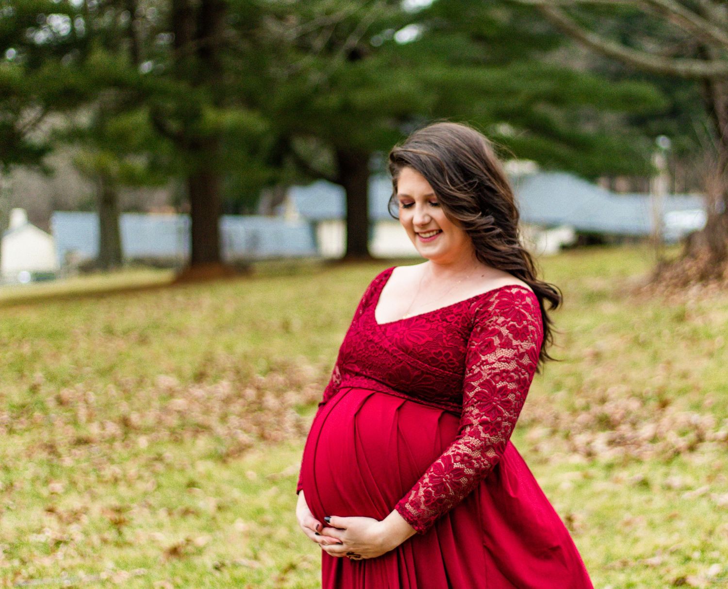 Woman in flowing red lace dress poses gracefully outdoors during autumn maternity photoshoot.