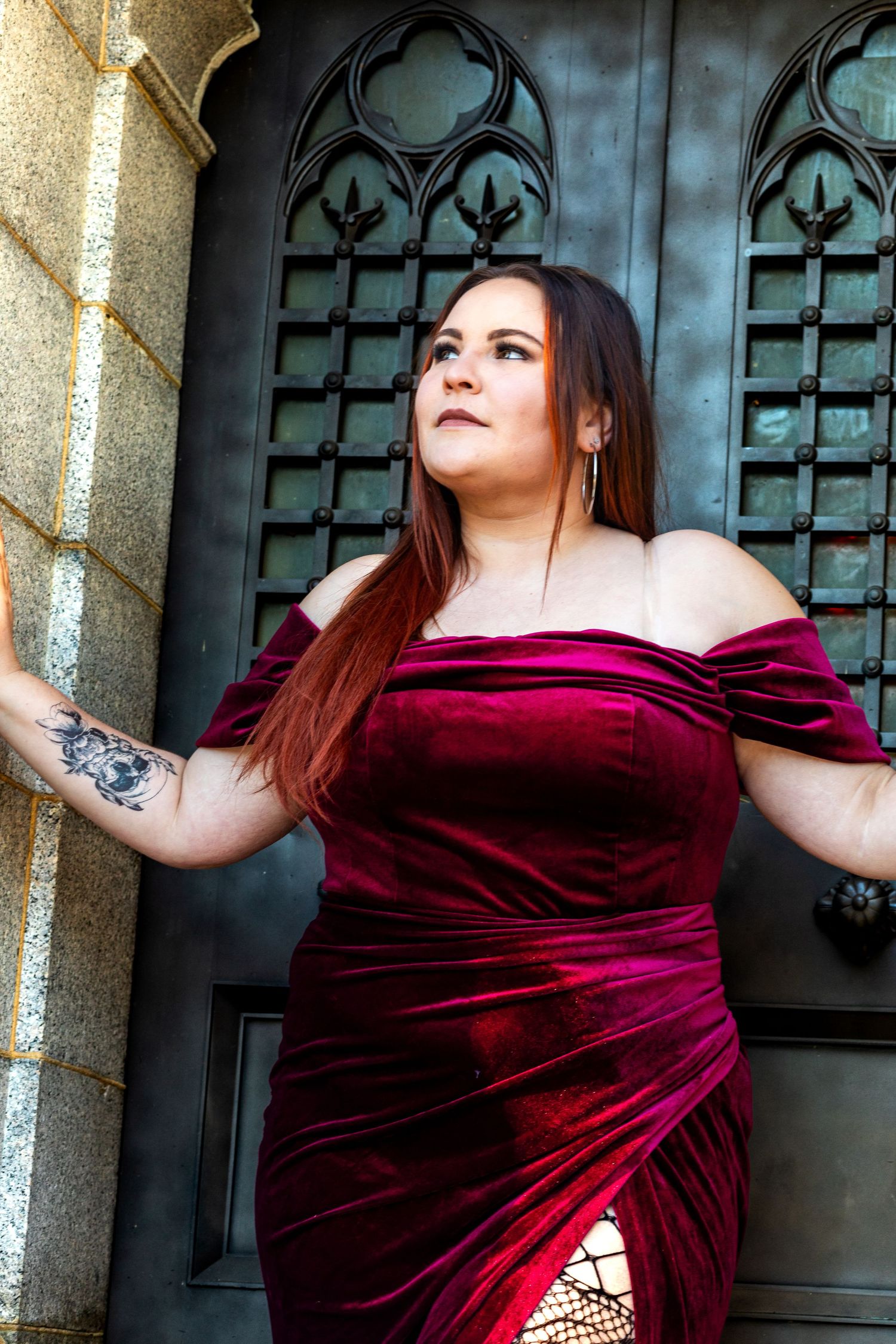 Woman in burgundy velvet off-shoulder dress poses against ornate gothic door with decorative windows.