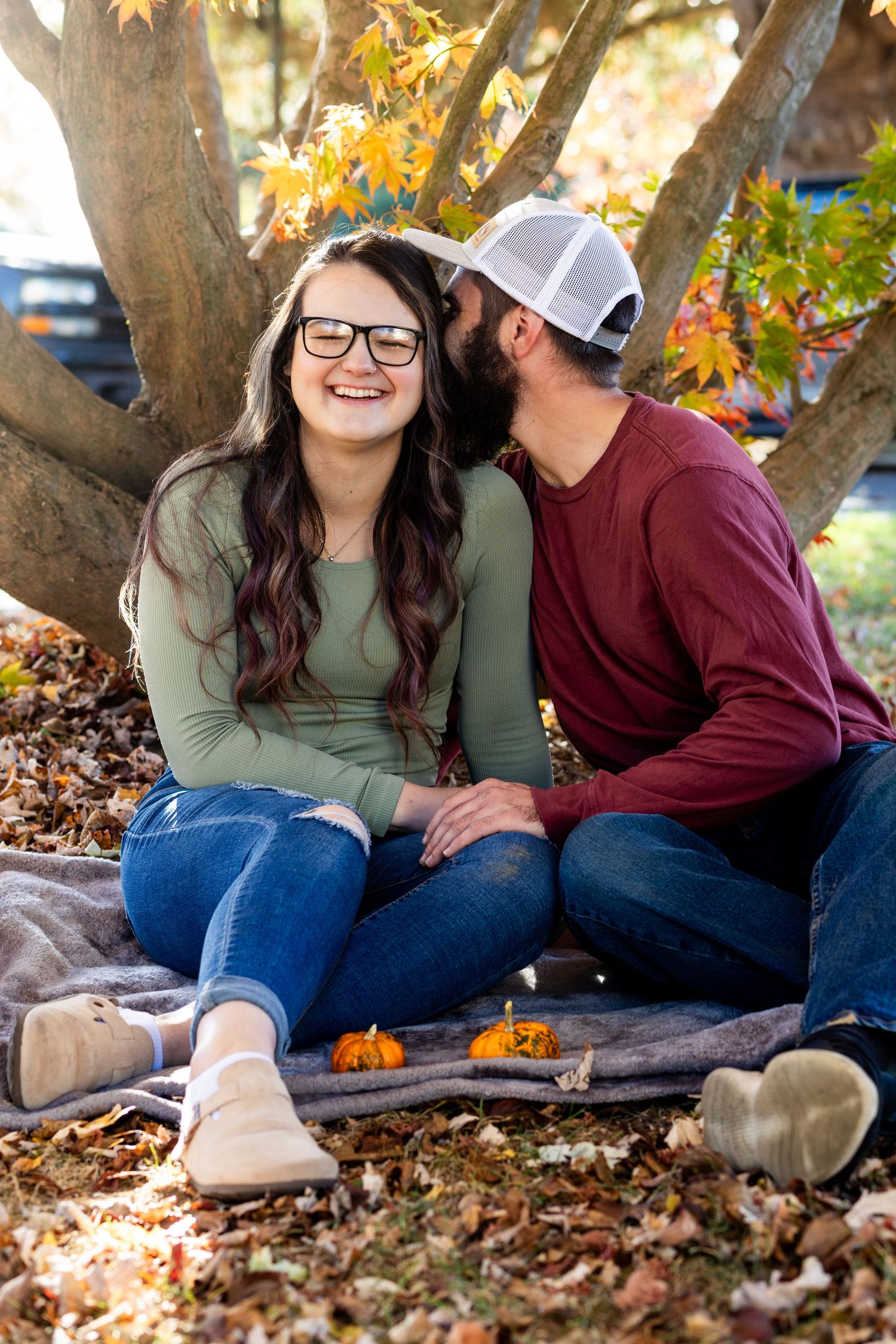 Couple sitting together on blanket surrounded by fall leaves under large tree.