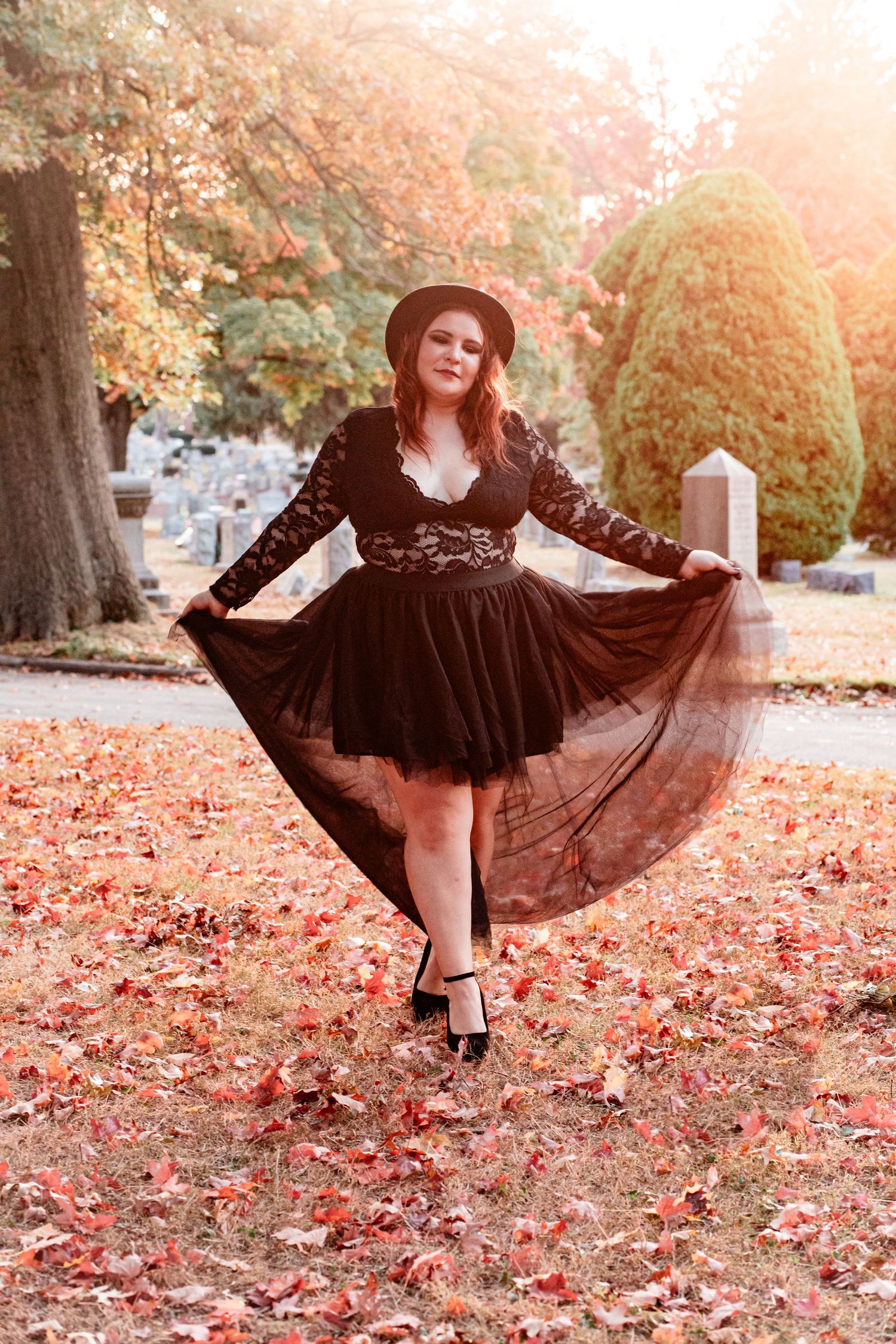 Person in black lace dress twirls among fallen autumn leaves during sunset photoshoot.
