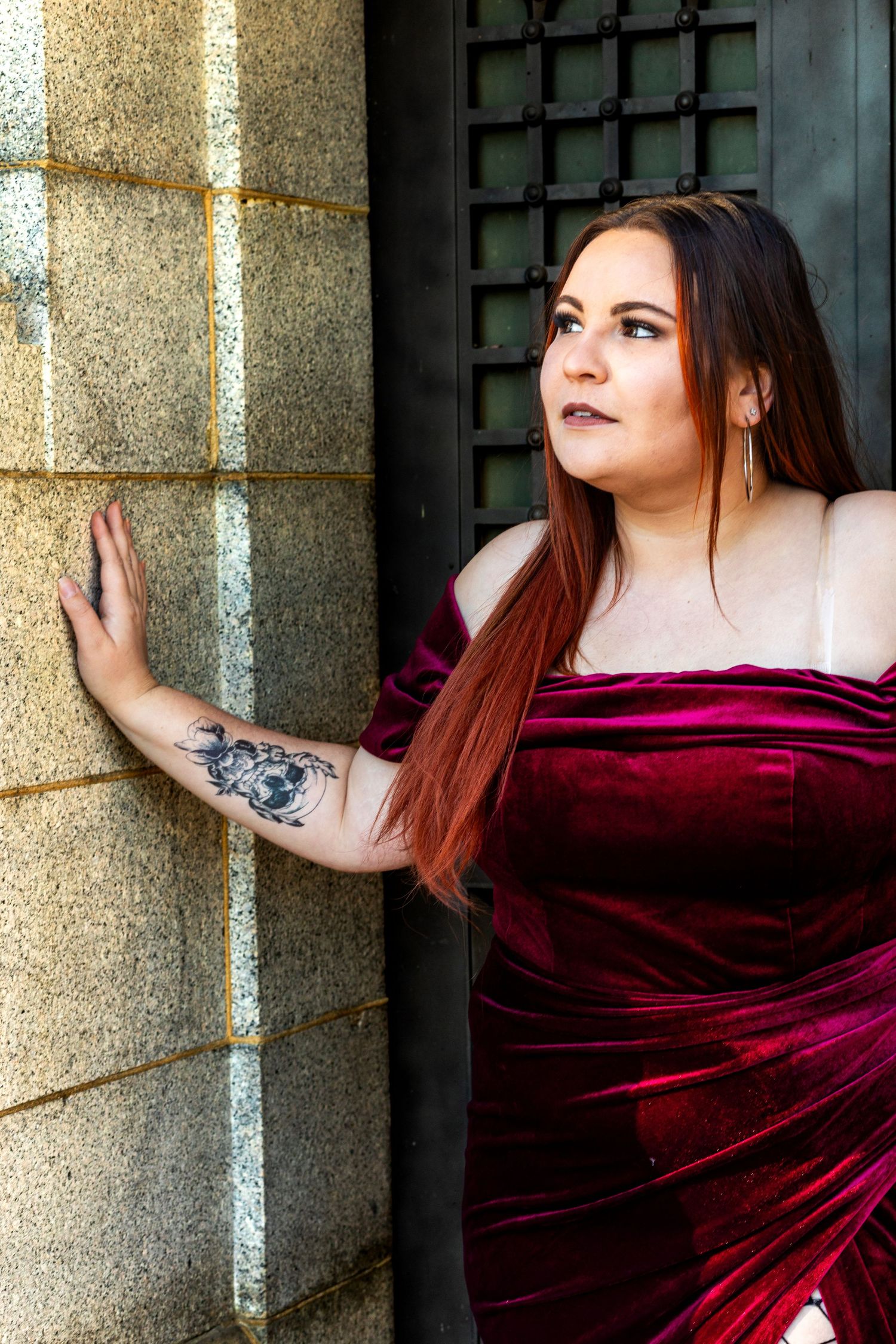 A woman in a burgundy velvet dress poses dramatically against a stone wall with decorative ironwork.