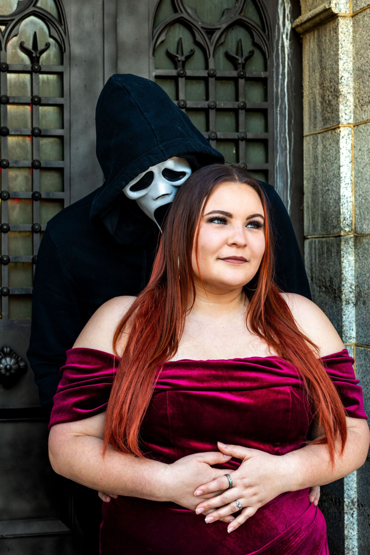 Figure in elegant burgundy dress poses with person wearing ghostface costume in gothic setting.