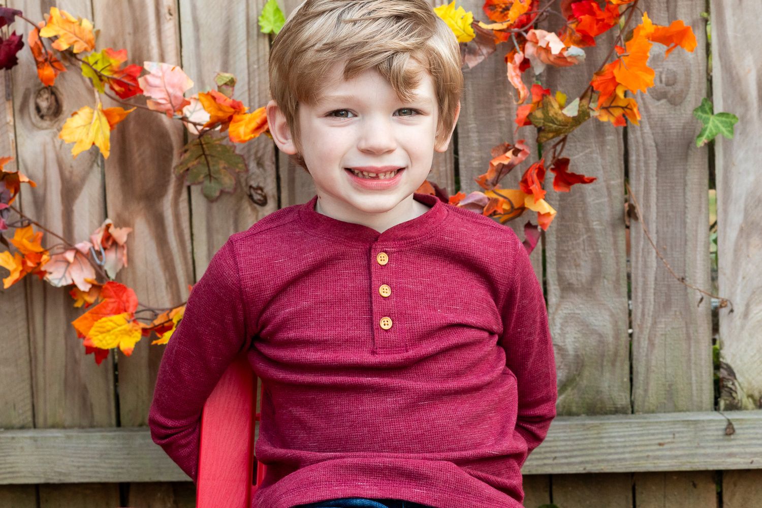 Child in burgundy henley shirt poses against wooden fence decorated with fall leaves.