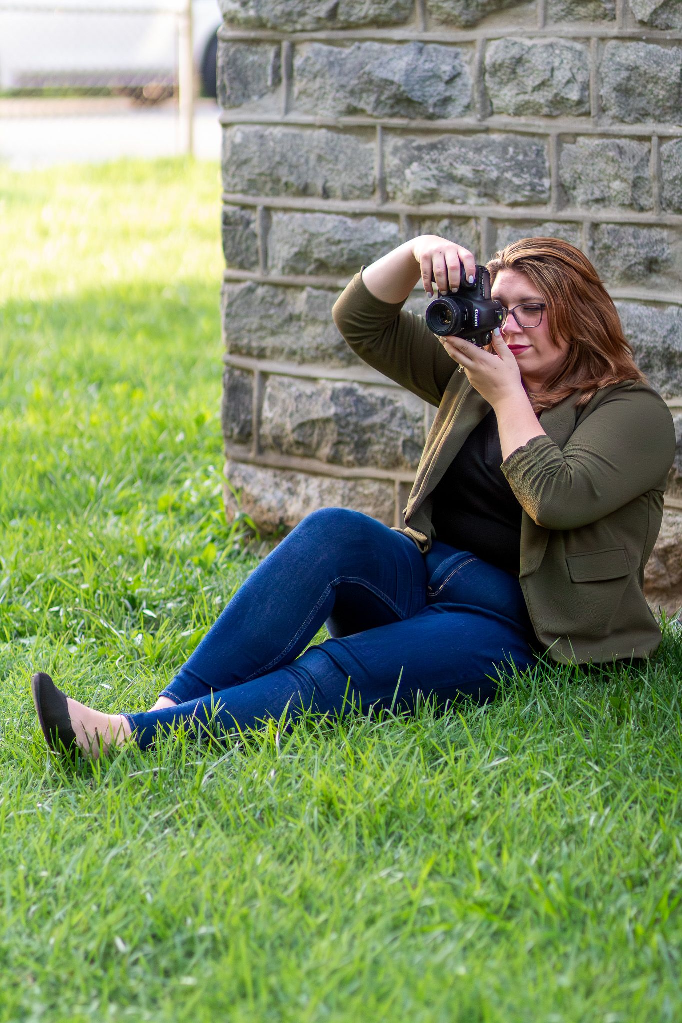 Photographer in casual attire taking photos while sitting on grass by stone wall.
