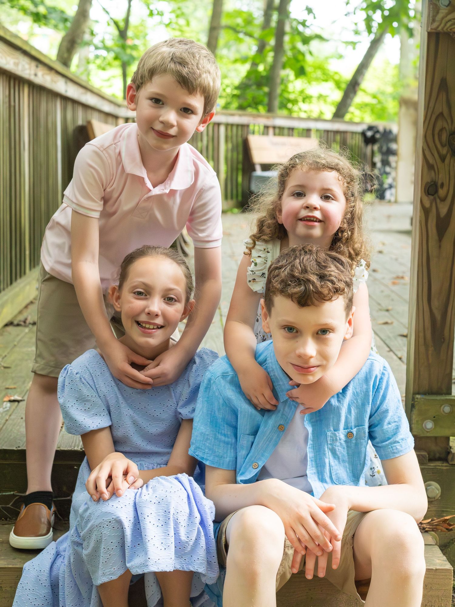 Four children pose together on wooden steps outdoors with greenery in the background.