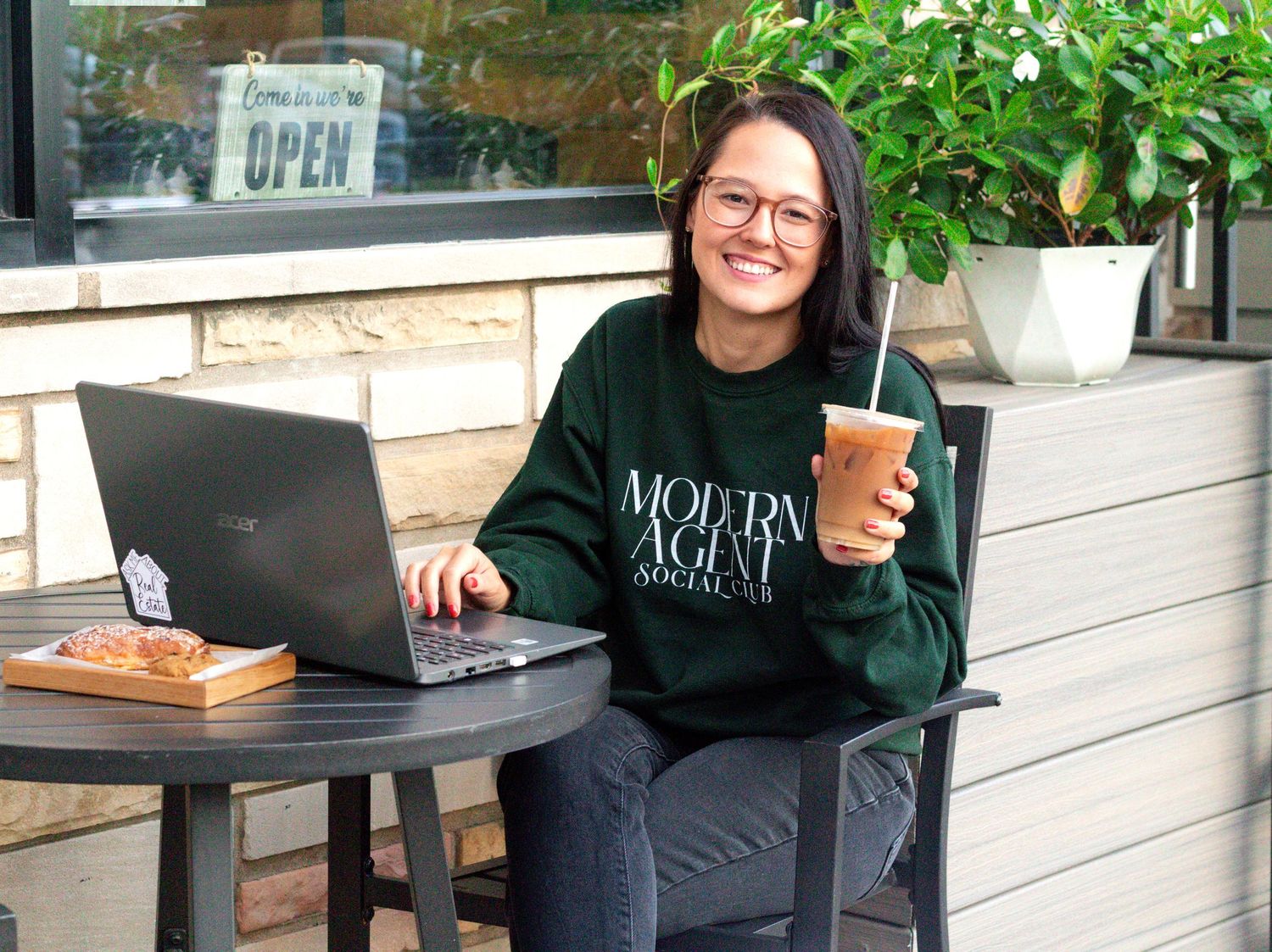 A laptop and drinks setup in an outdoor cafe seating area with wooden tables and lush greenery.