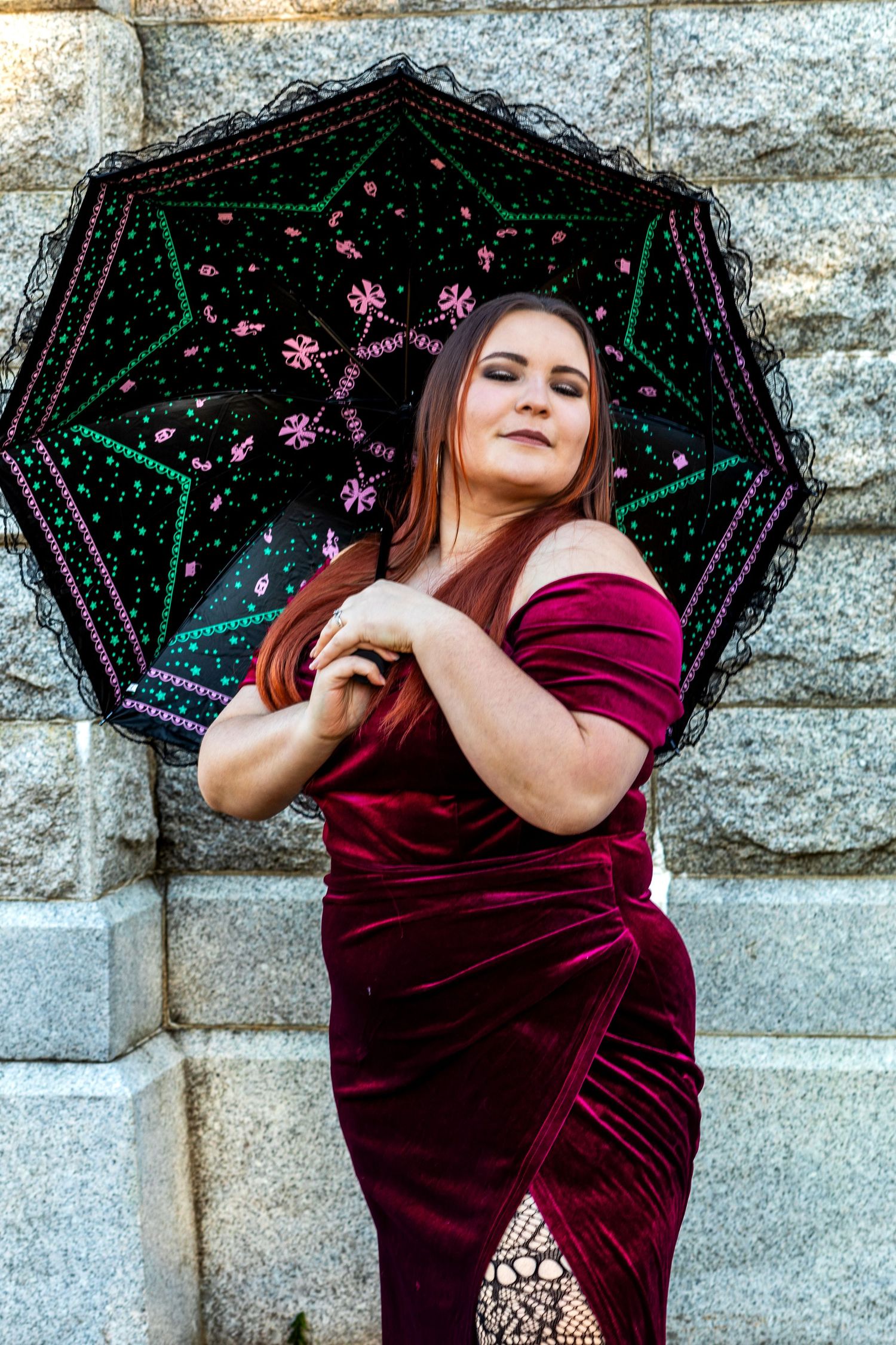 Woman in burgundy velvet dress poses with decorative black umbrella with pink floral designs against stone wall.