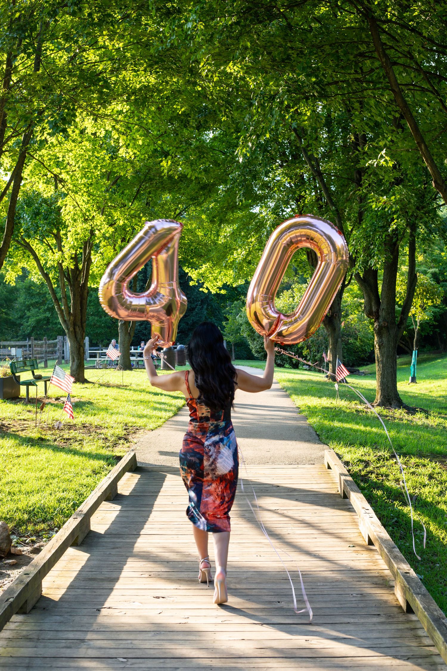 Person holding metallic number 40 balloons while walking on wooden boardwalk surrounded by green trees.