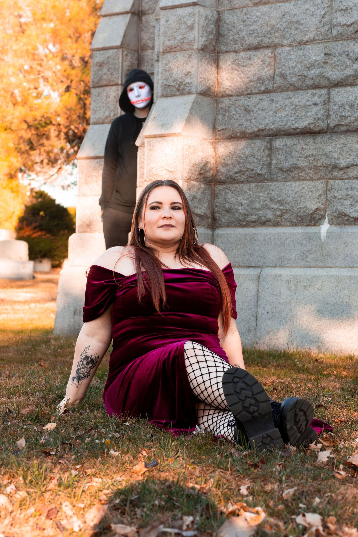 Someone in a burgundy velvet dress with fishnet stockings poses near stone wall while another figure stands in background.