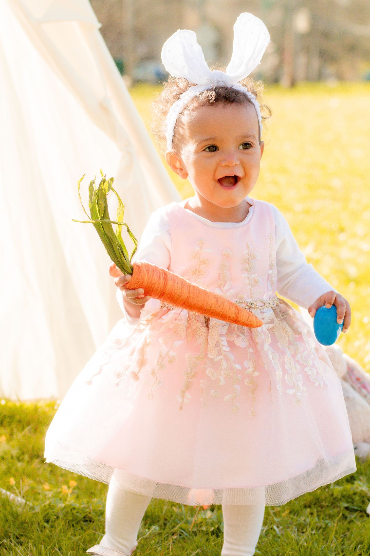 Baby in white dress and bunny ears holding a large carrot in sunny garden setting.