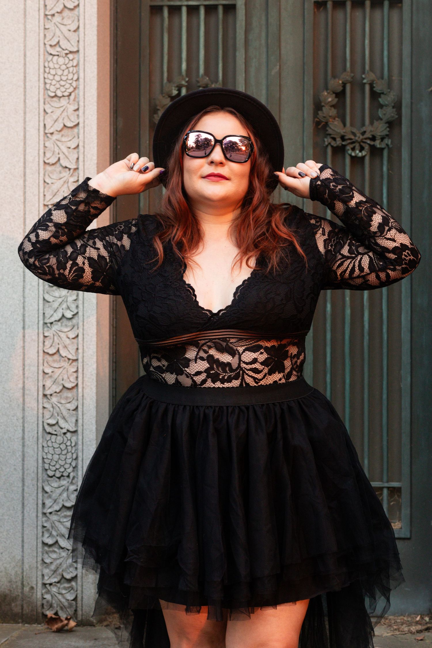 Woman in a black lace mini dress strikes playful poses against a stone wall backdrop while wearing sunglasses.