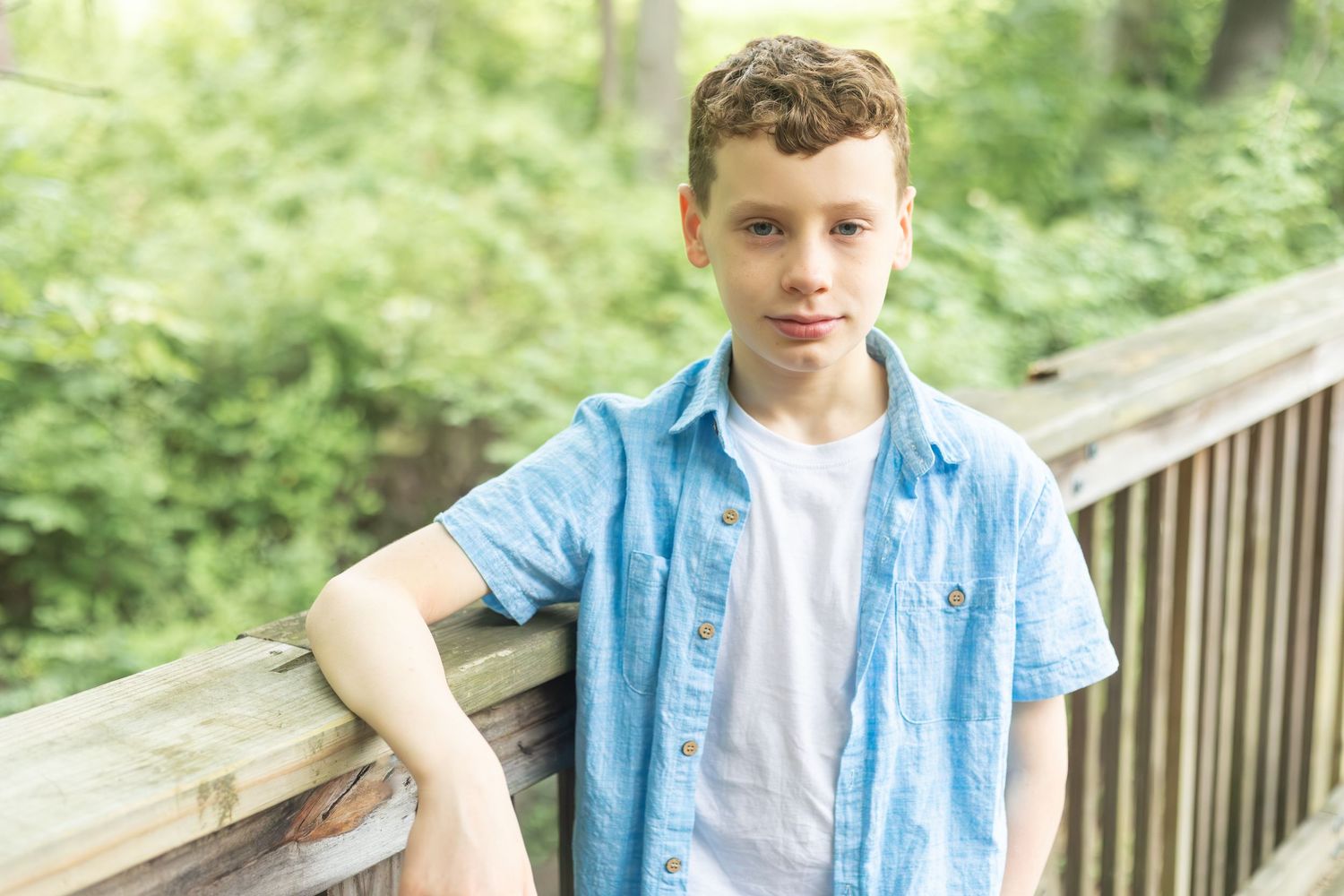 A youth wearing a light blue button-down shirt over a white t-shirt leans against a wooden railing in a wooded setting.