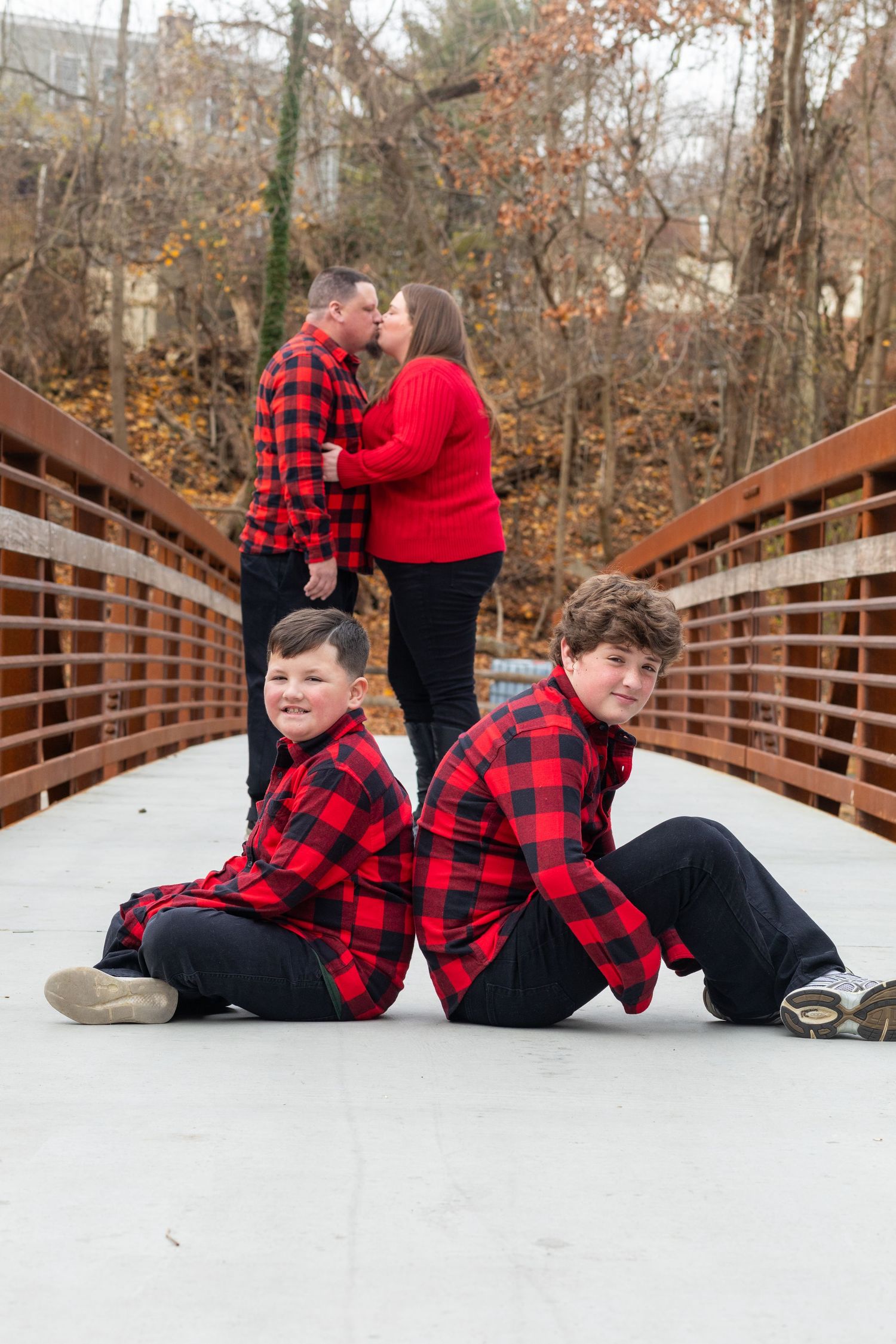 Family wearing coordinating red and black plaid shirts pose together on wooden bridge.