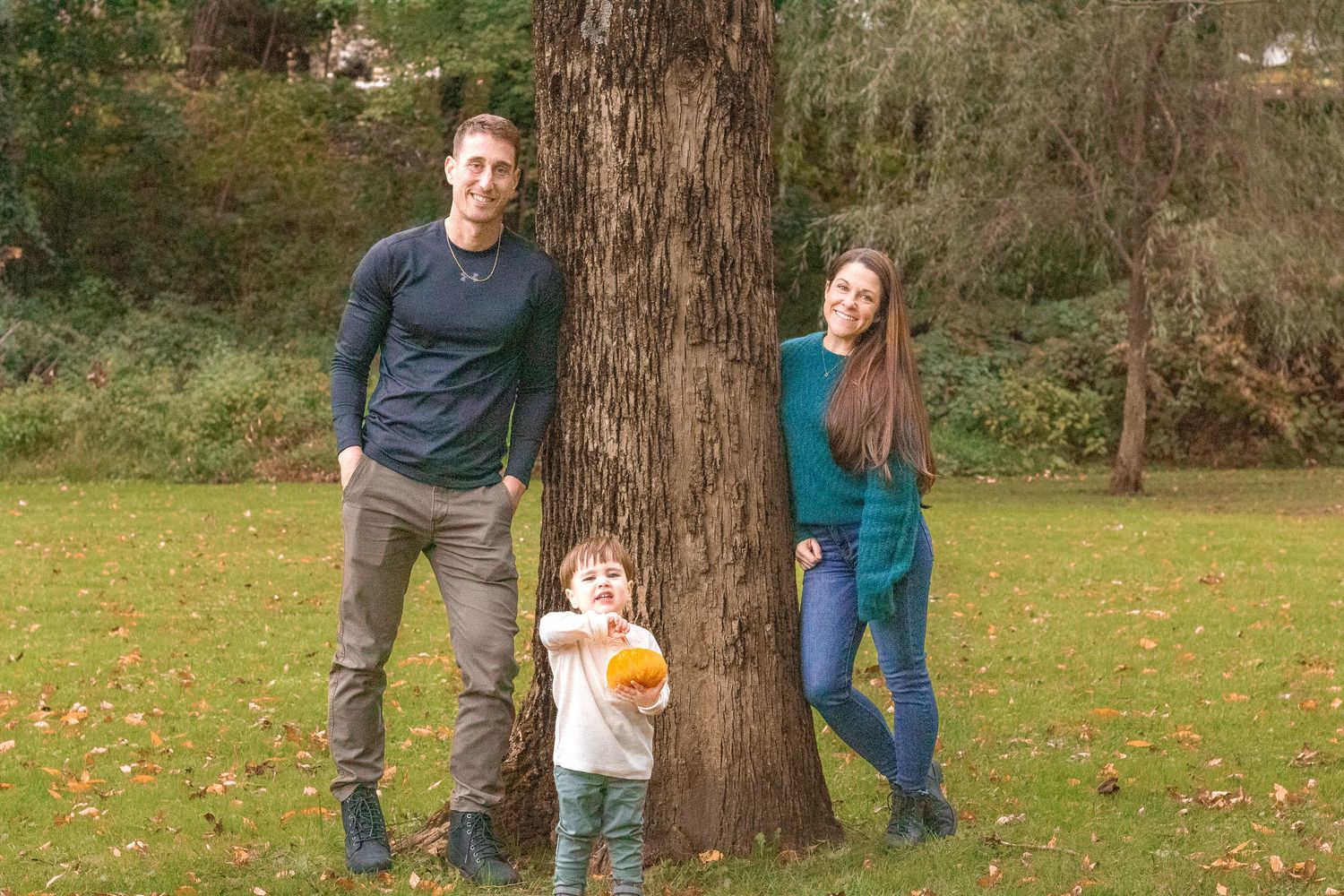 Family walking and playing around large tree trunk in autumn park setting.