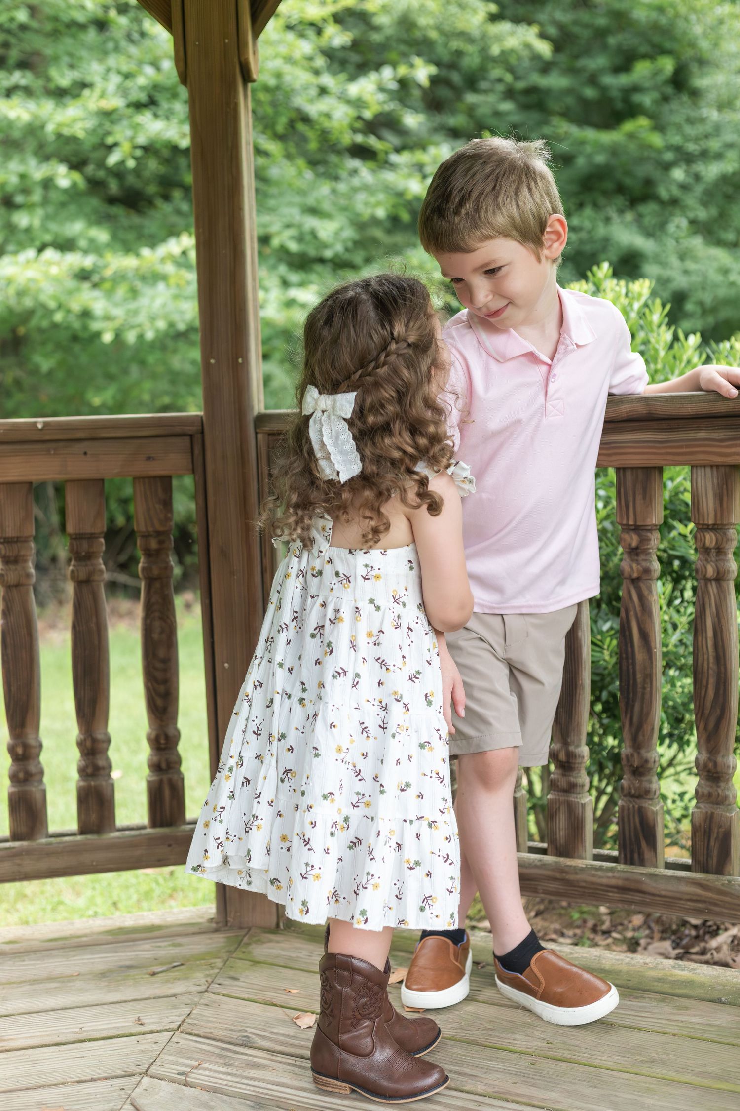 On a wooden deck, a small child in a floral dress stands close to someone in a pink shirt looking downward.