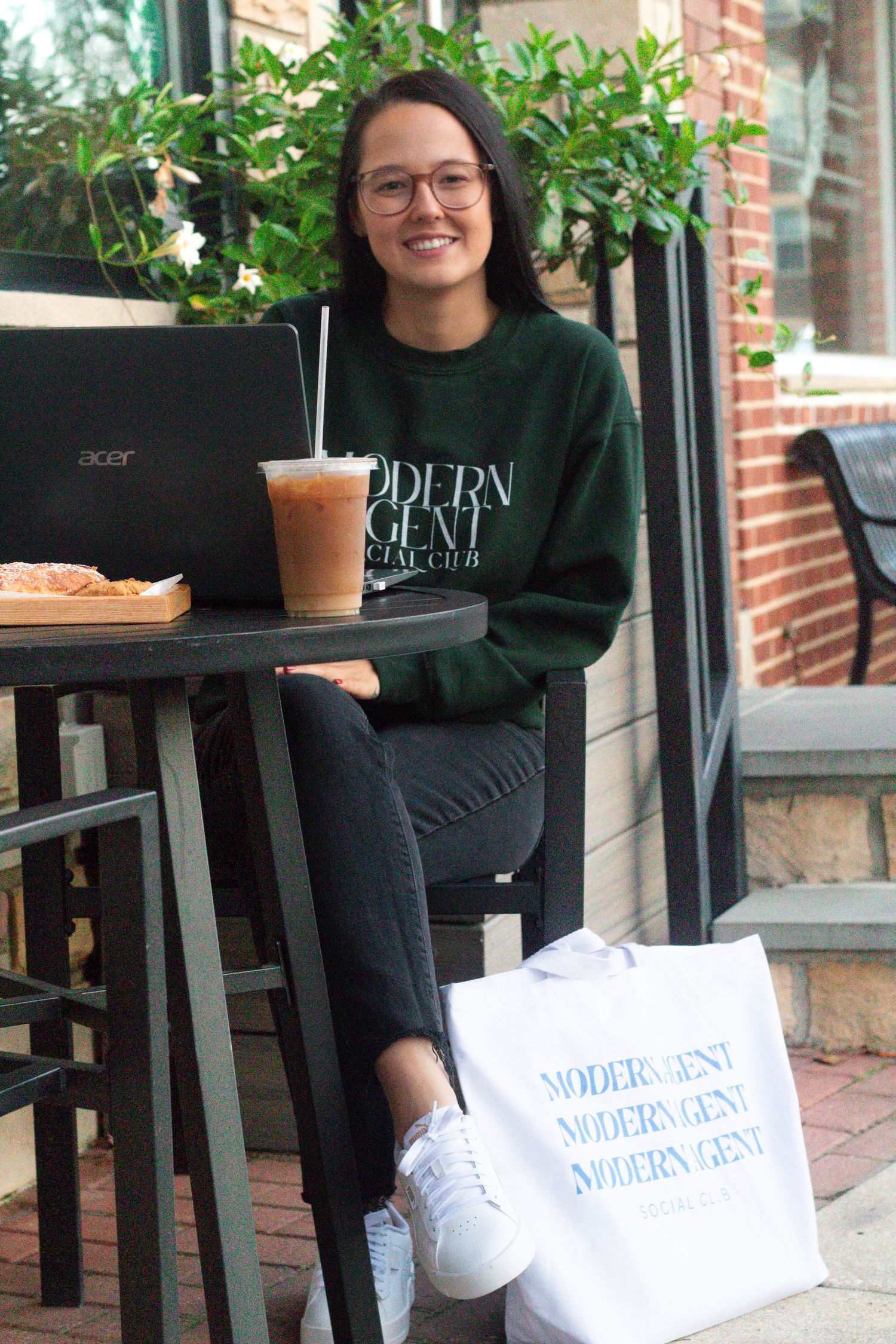 Student working on laptop at outdoor cafe with iced coffee and shopping bag.