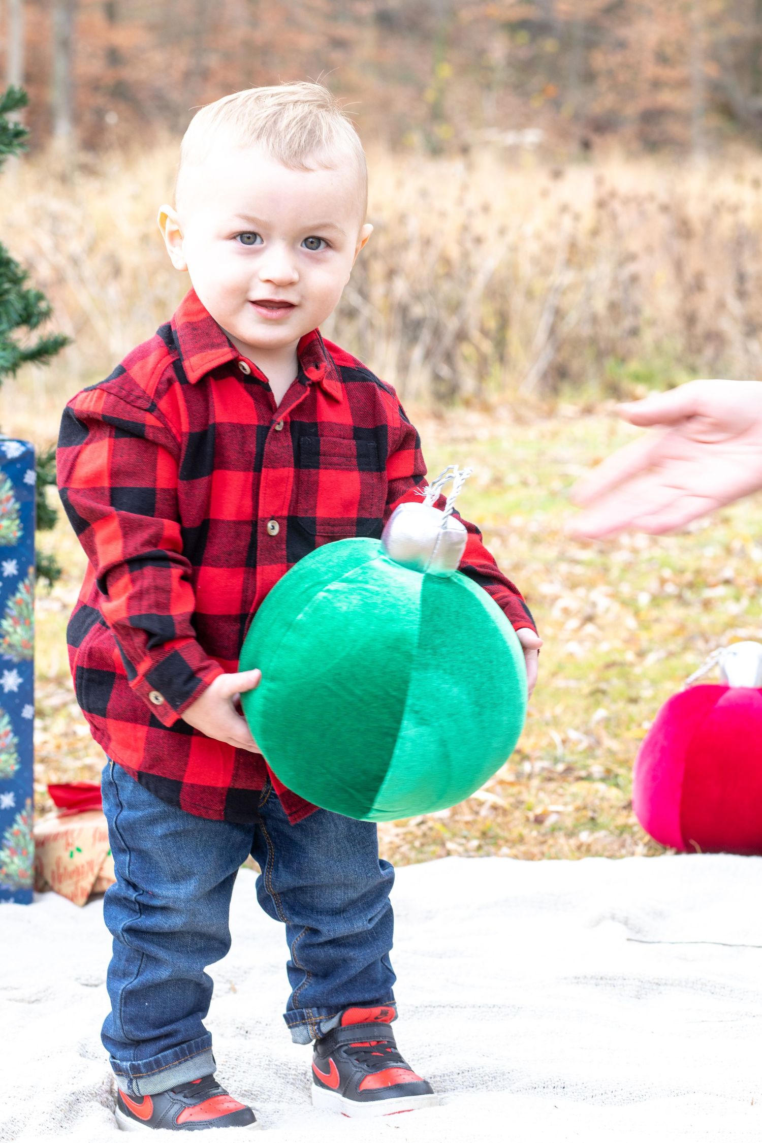 Toddler in red plaid flannel and jeans holds large green ornament outdoors.