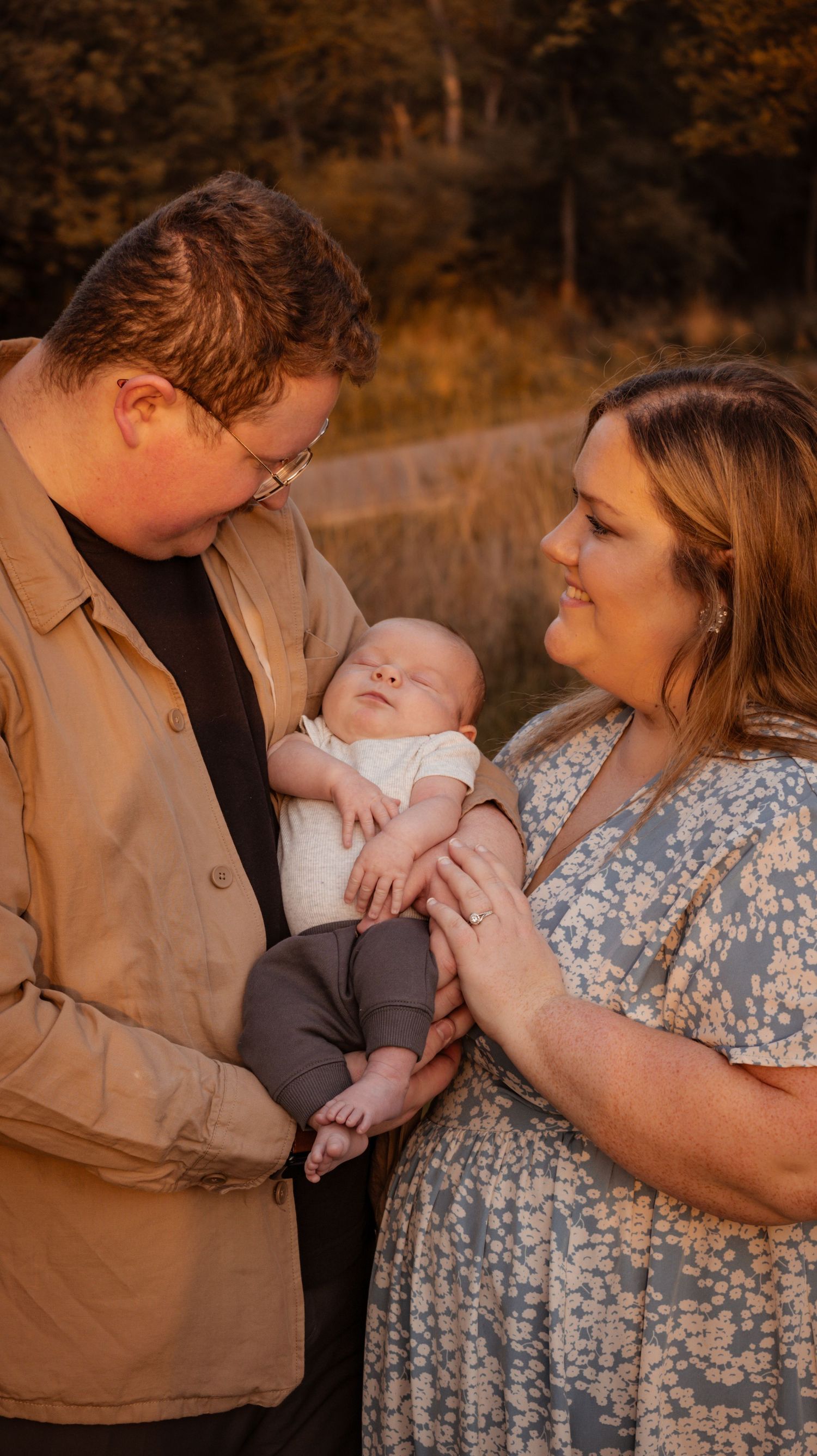 A mom and dad smiling sweetly as they hold their newborn baby in front of a field