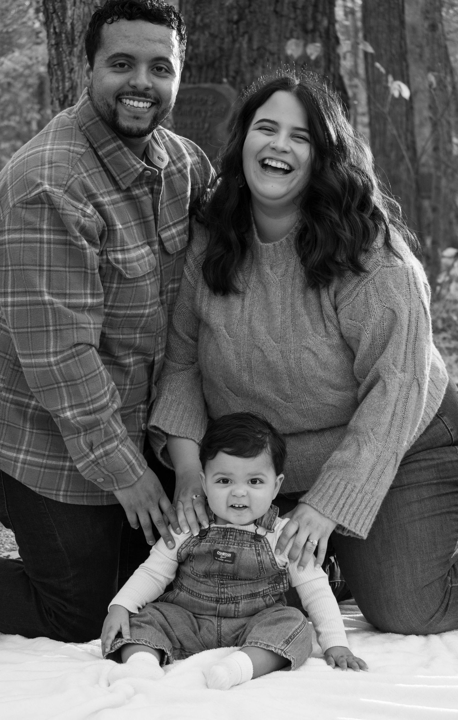 A black and white photo of a mom and dad smiling while trying to get their little boy to sit still with them on the ground