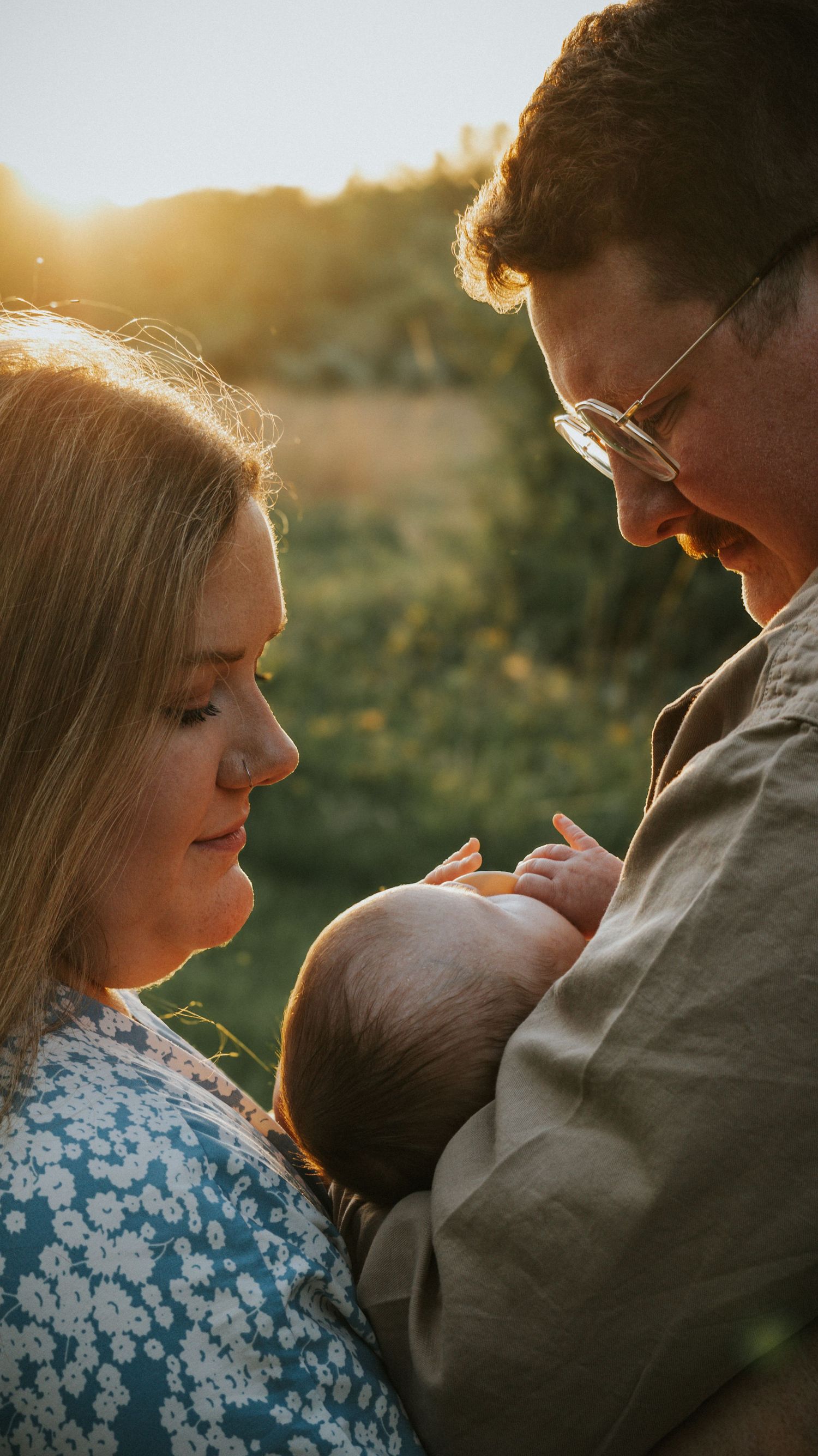 A mom and dad smiling sweetly as they hold their newborn baby in front of a field while the sun is coming through