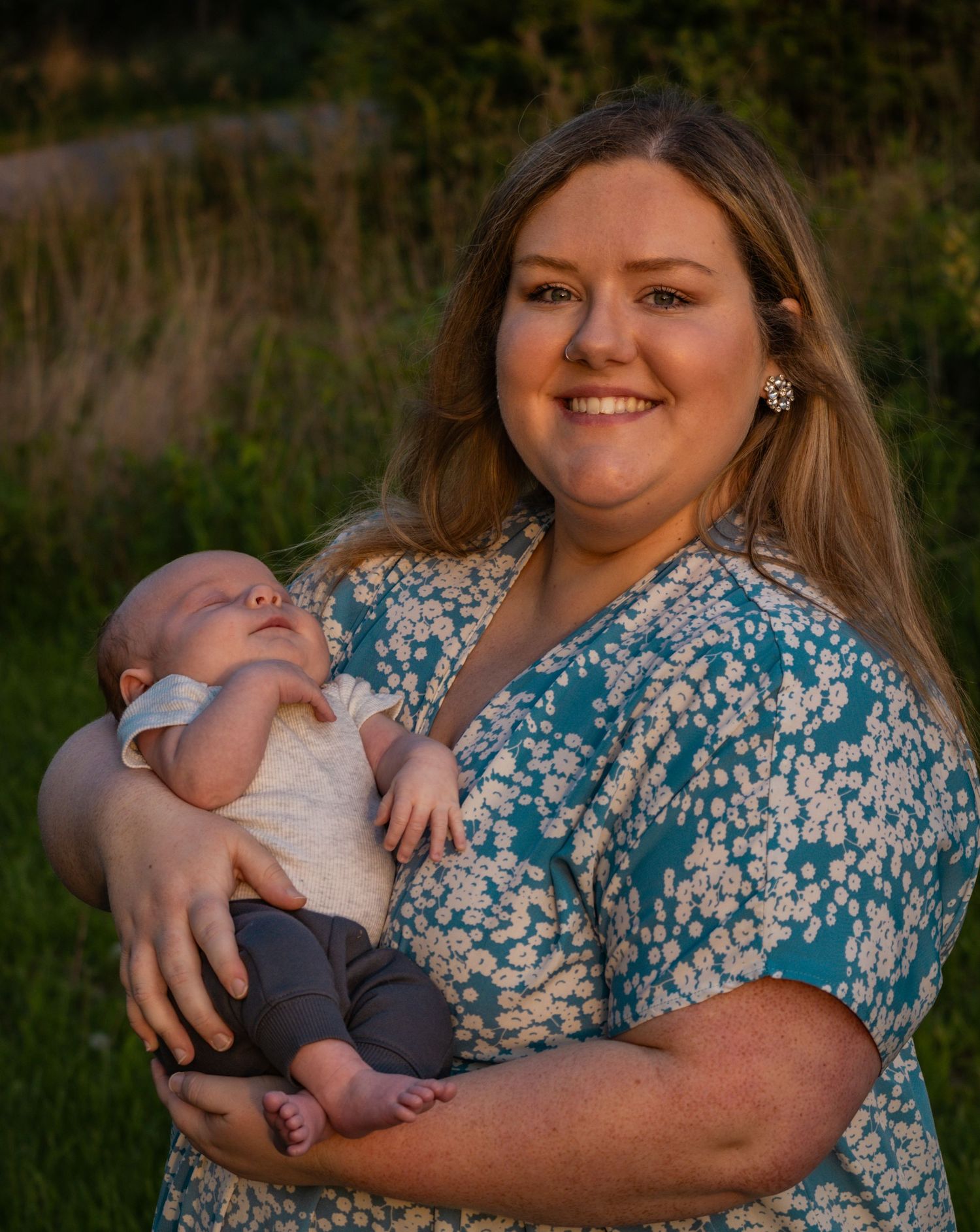 A smiling mom wearing a blue sundress holds her newborn baby by a field
