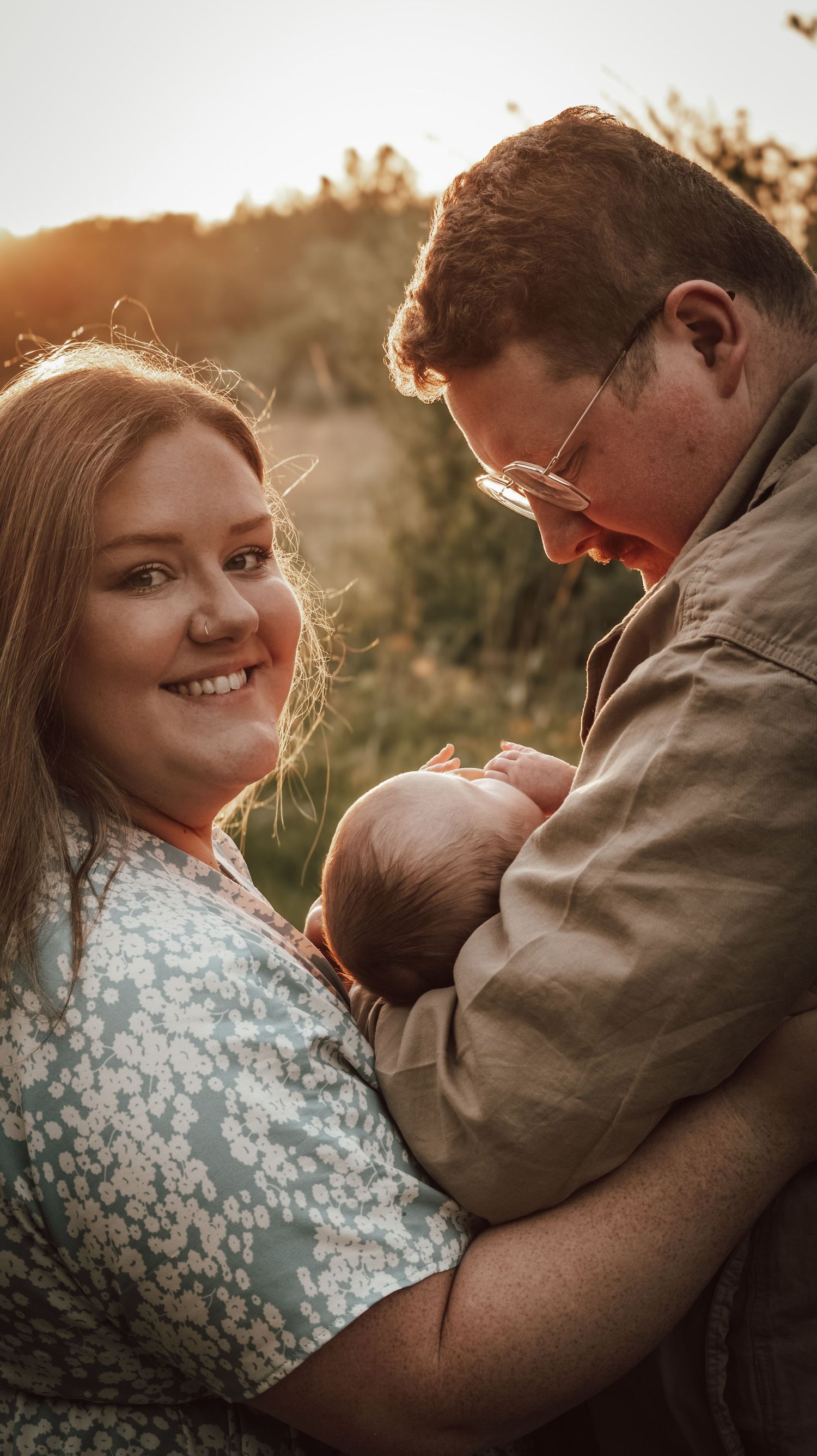 A mom and dad smiling sweetly as they hold their newborn baby in front of a field while the sun is coming through