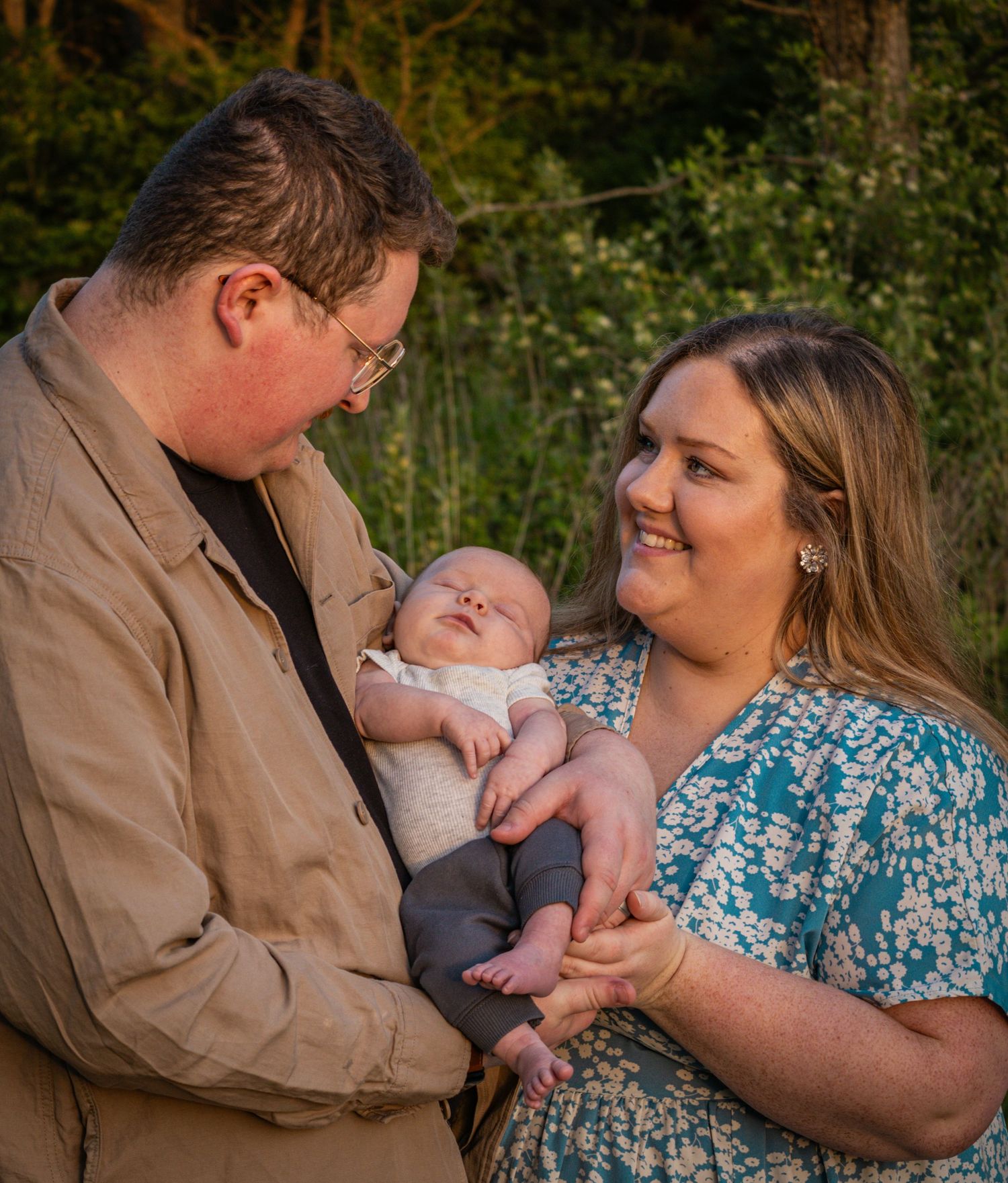 A mom and dad smiling sweetly as they hold their newborn baby in front of a field