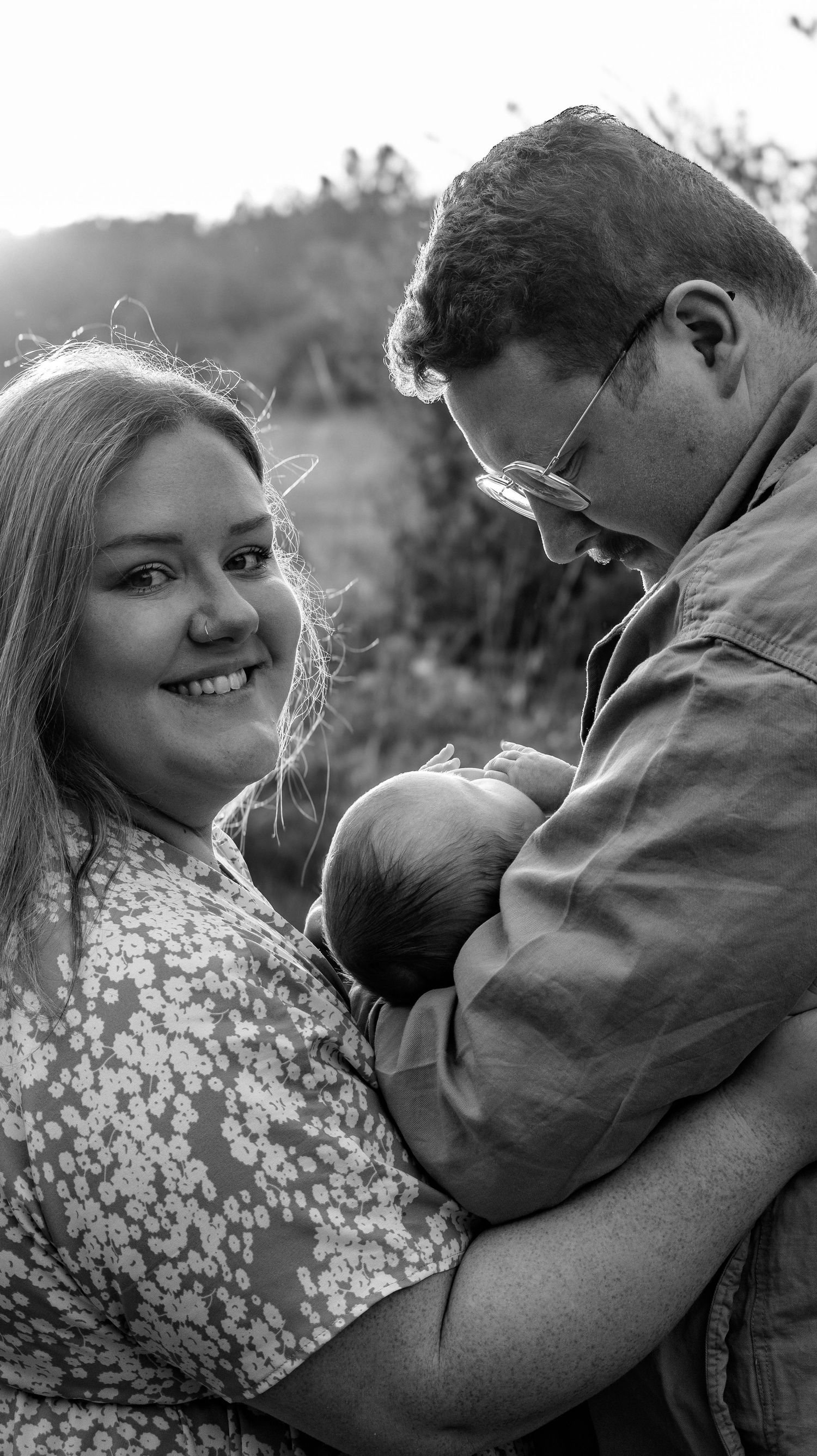 A black and white photo of a mom and dad smiling sweetly as they hold their newborn baby in front of a field