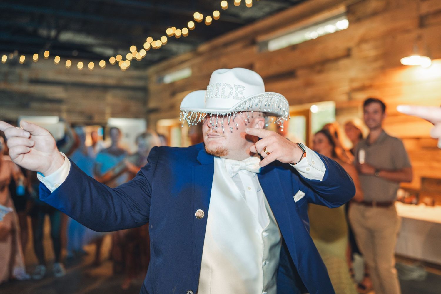 Groom in navy suit and white cowboy hat dancing joyfully at rustic wedding reception with string lights.