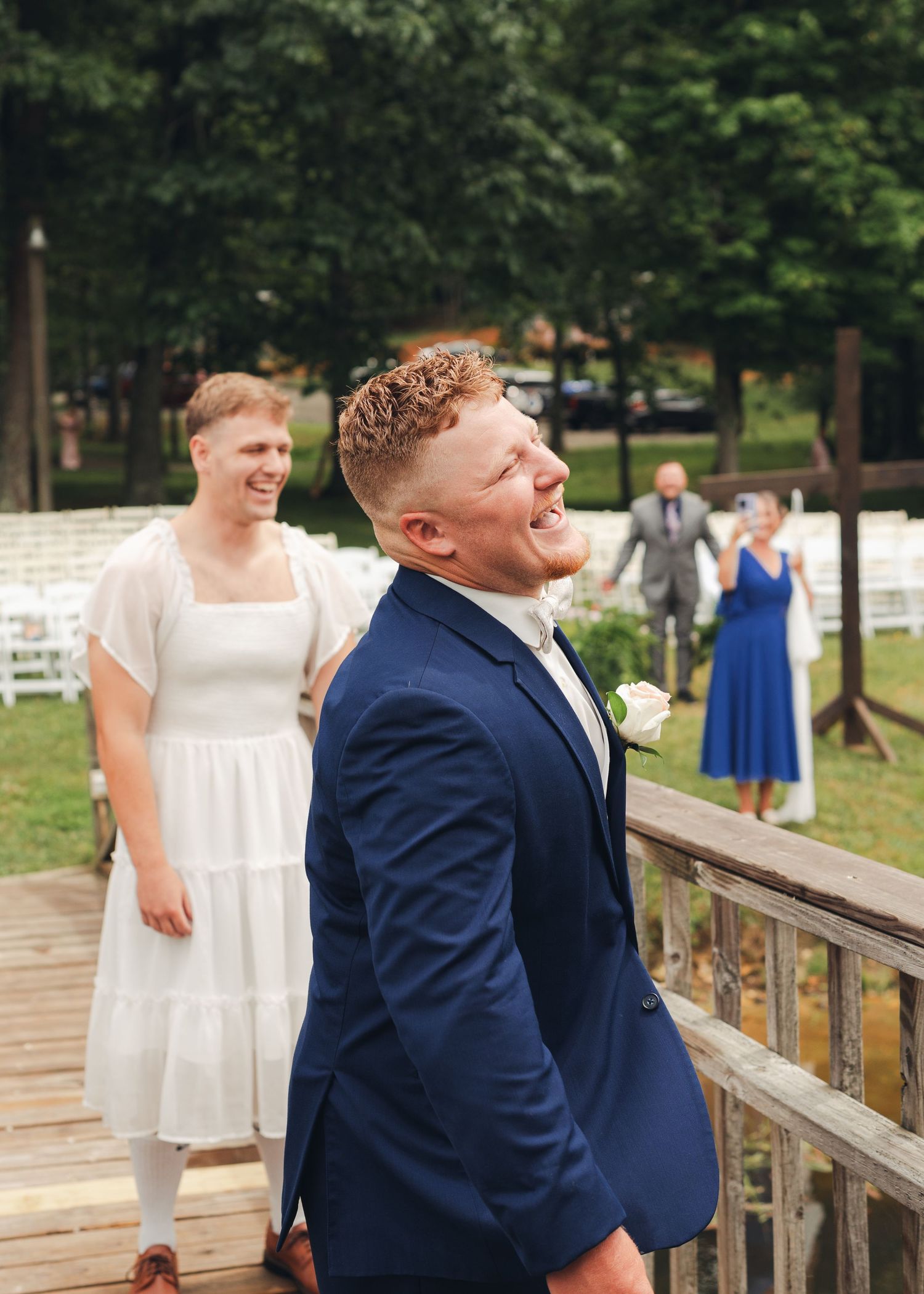 Groom in navy suit smiling joyfully at outdoor wedding ceremony with guests in background.