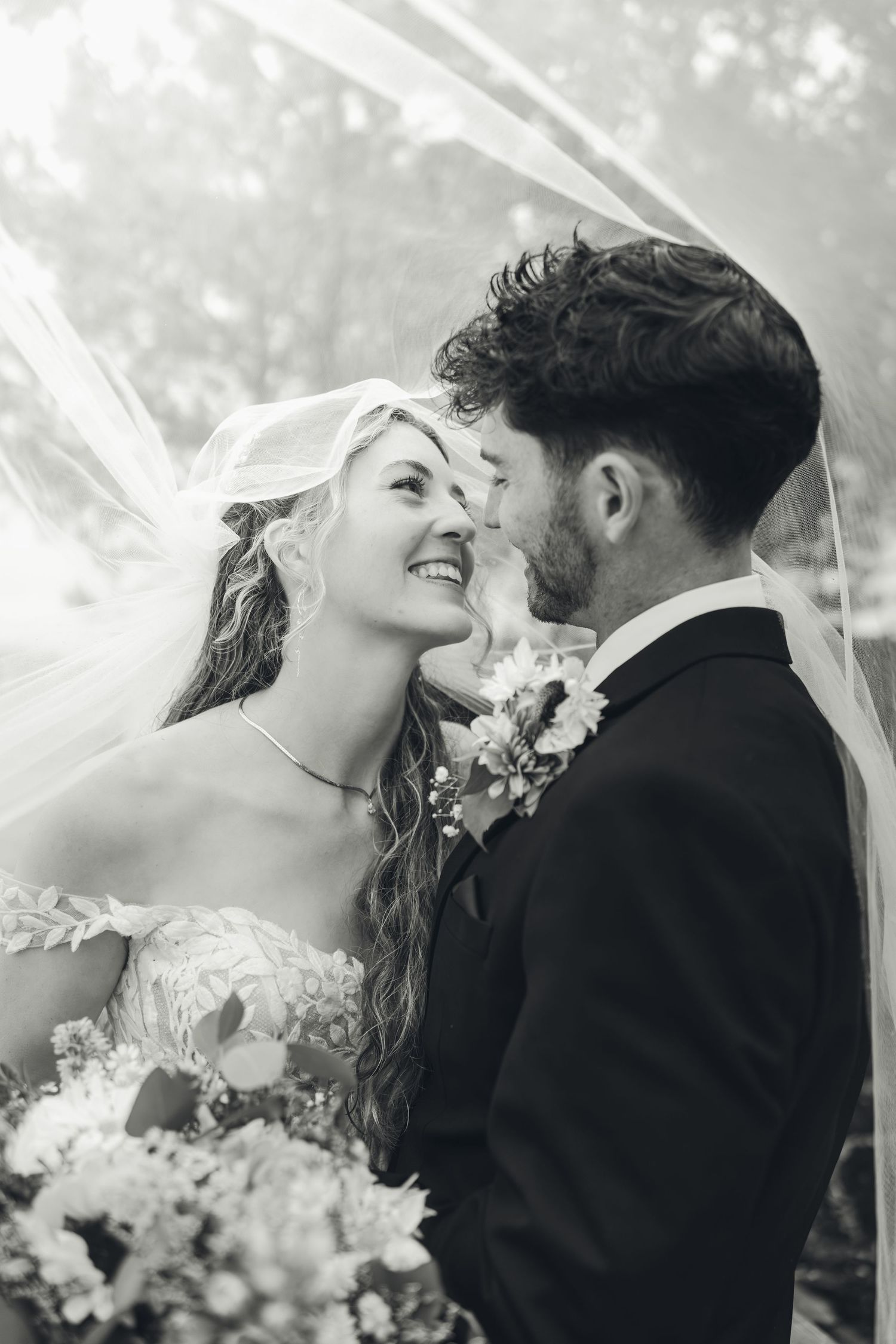 Black and white photo of a smiling bride and groom sharing a romantic moment under a flowing wedding veil.