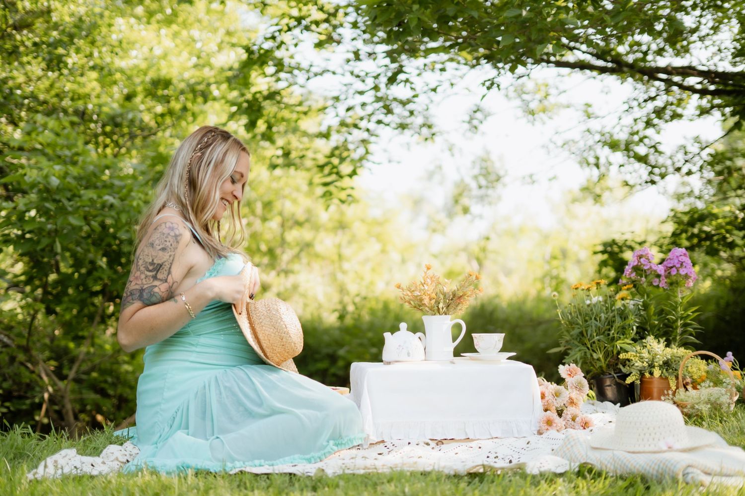 Tattooed pregnant woman in mint dress enjoying outdoor picnic with tea set on white blanket in garden.