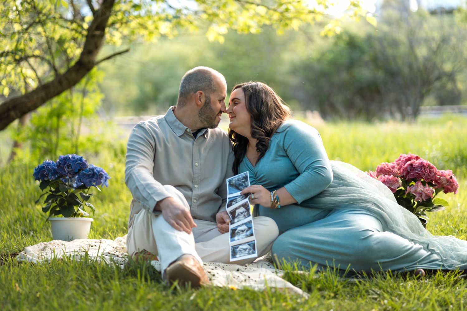 Couple sharing a kiss during outdoor maternity session with ultrasound photos and flowers on a blanket.