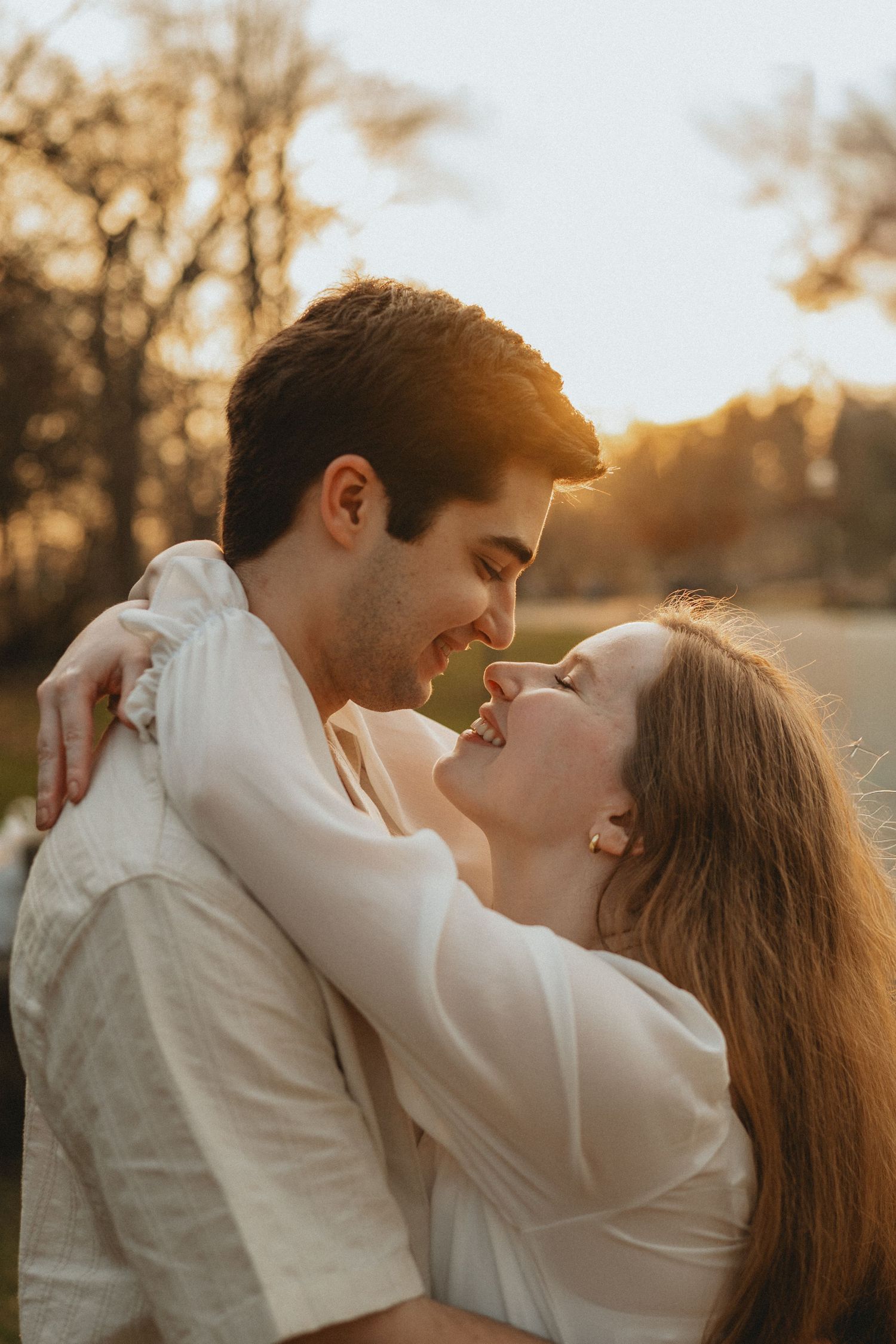 Smiling couple embracing nose-to-nose during warm golden sunset engagement session in an outdoor park setting.
