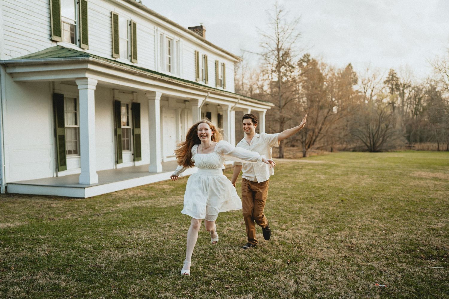 A joyful couple runs hand in hand across a green lawn in front of a classic white historic house.