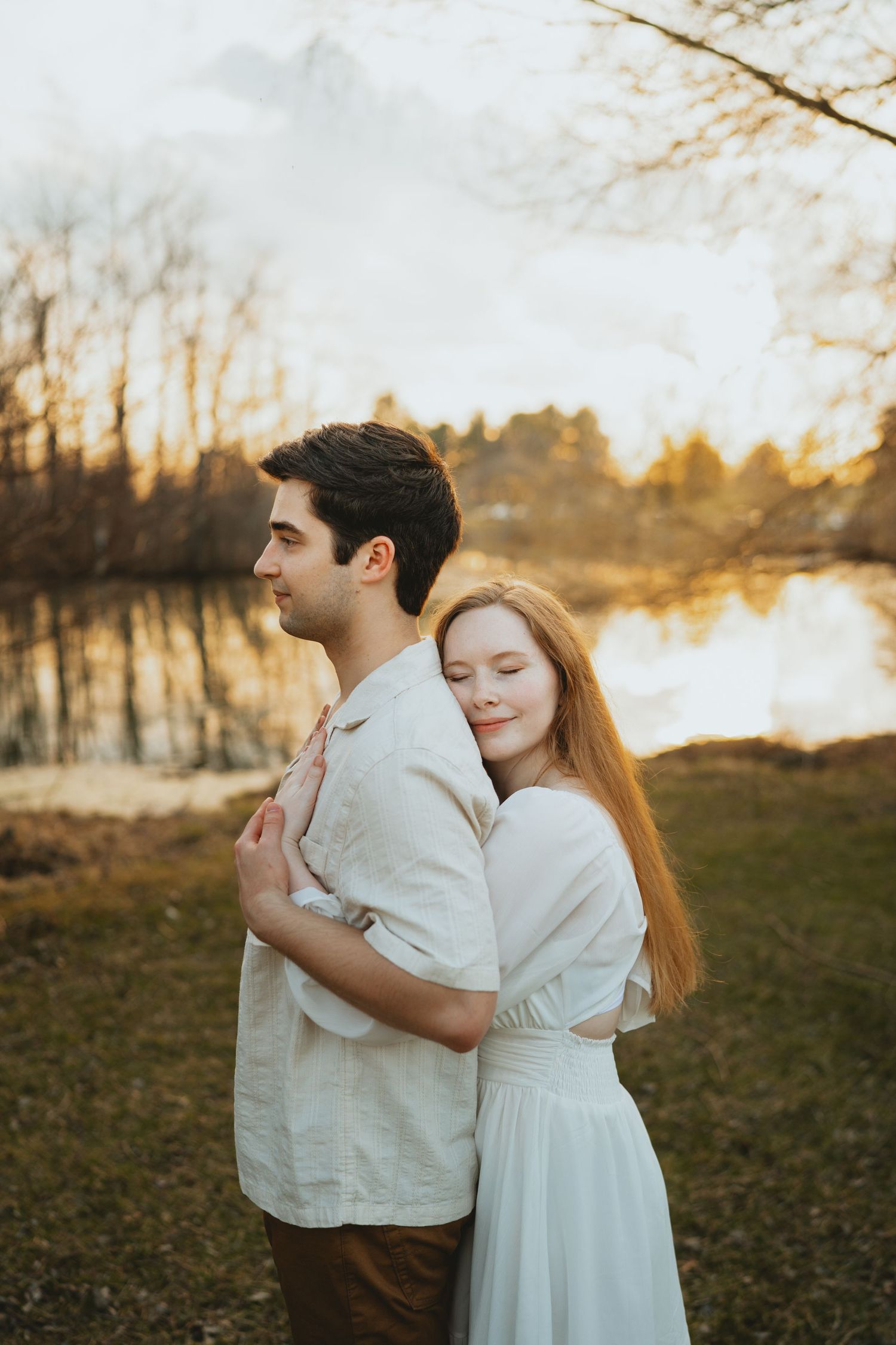 Woman in white dress embraces man from behind near a reflective lake at golden hour.