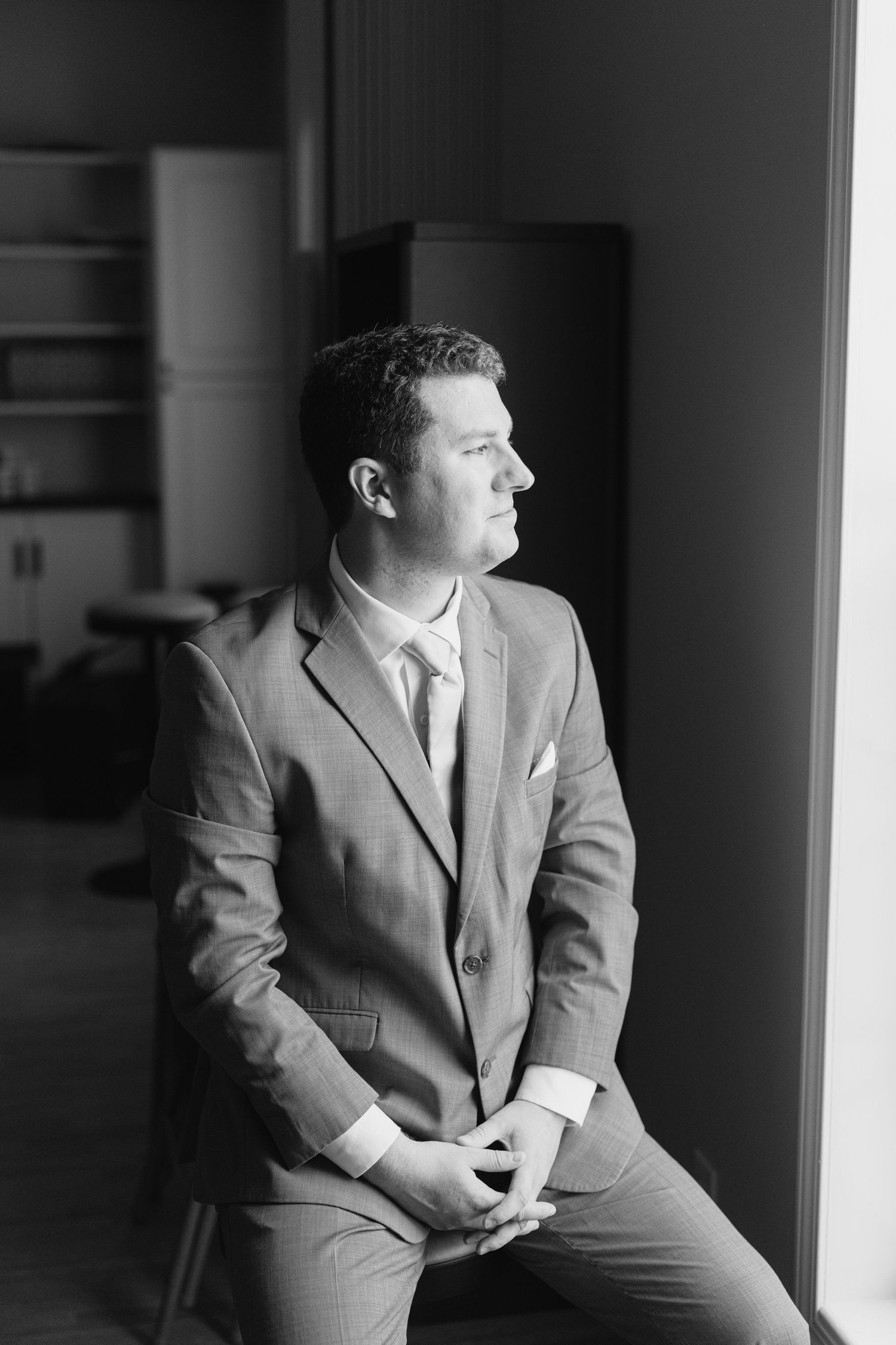 Black and white portrait of a groom in a suit sitting by a window, gazing thoughtfully to the side.