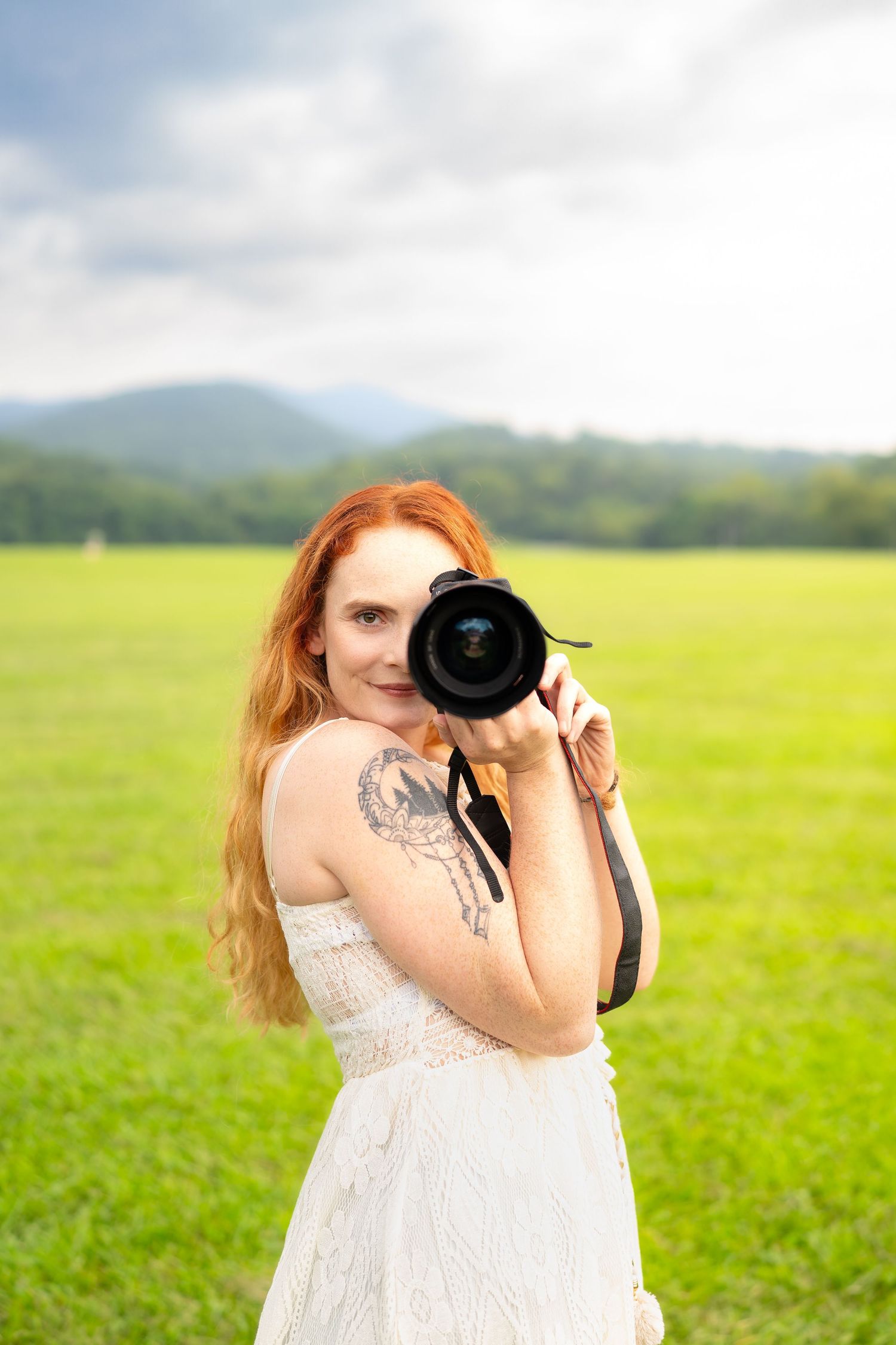 Red-haired woman in white dress holding a camera up to photograph in a scenic green field.