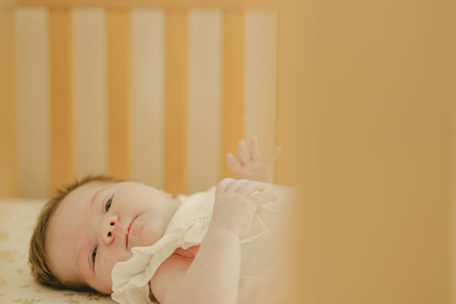 Awake newborn baby girl lying in wooden crib looking up with hand raised in warm light.