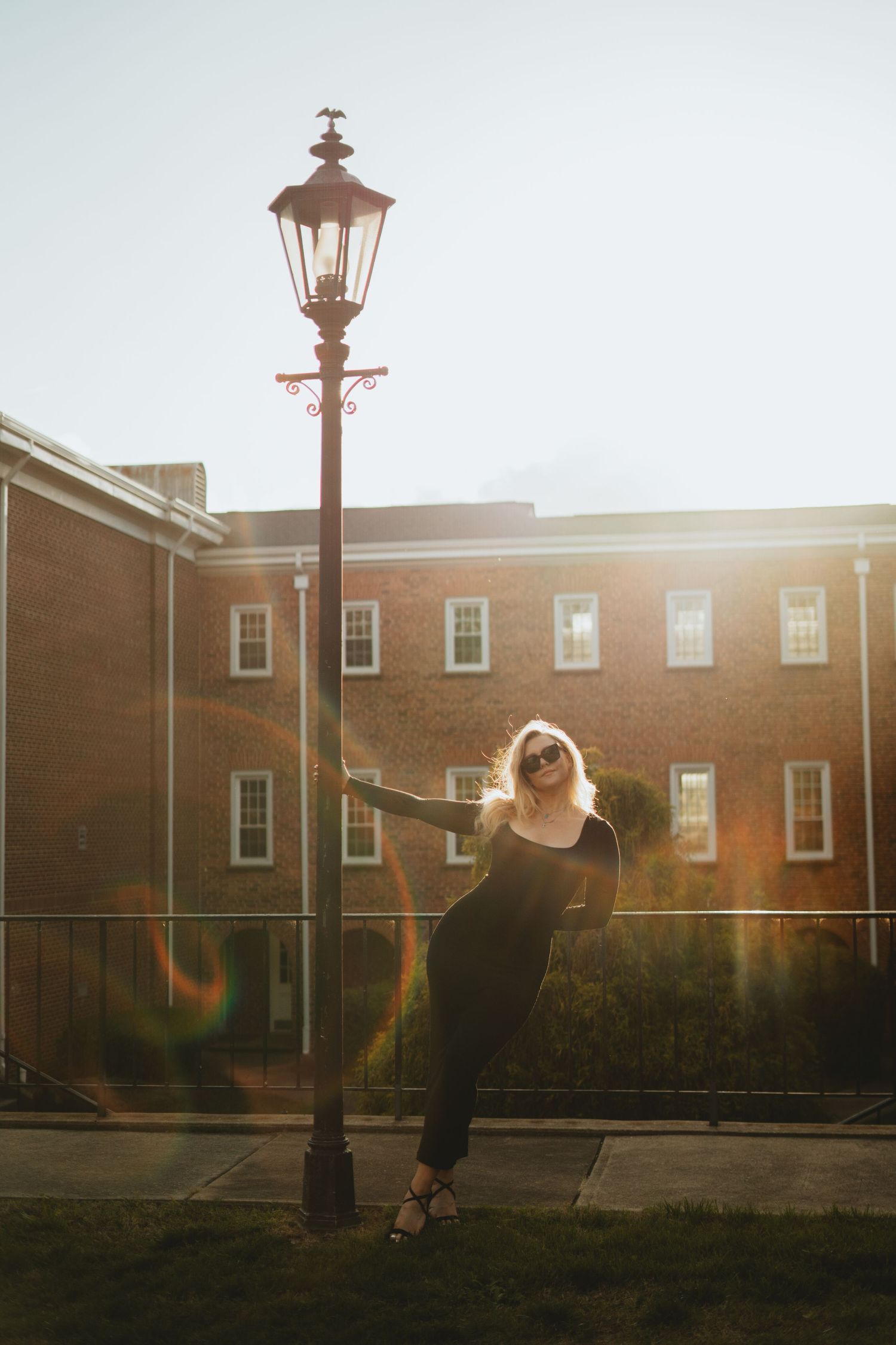 Blonde woman in black outfit leaning against a lamp post in front of a brick building at sunset.