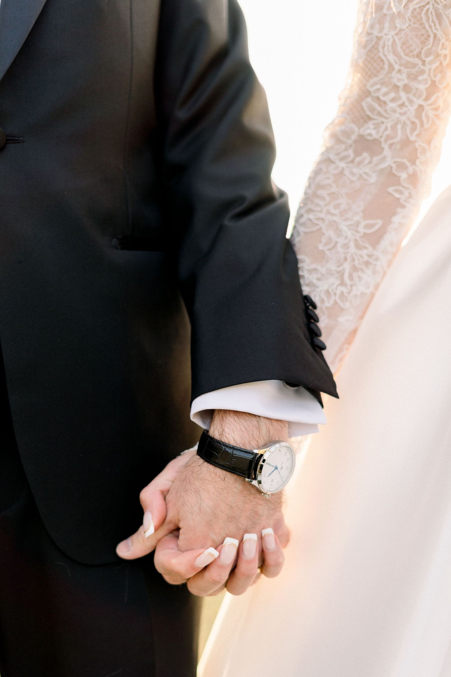 Close-up of bride and groom holding hands, groom wearing black watch, bride in lace sleeve wedding dress.