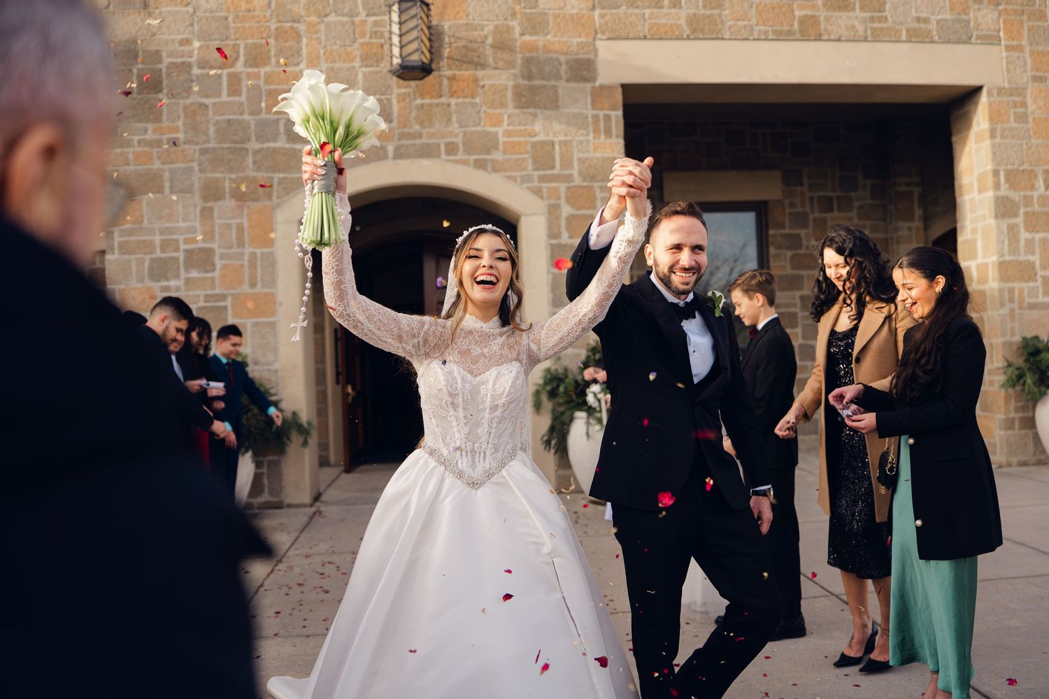 Happy bride and groom celebrate wedding exit with raised hands amid falling rose petals outside stone venue.